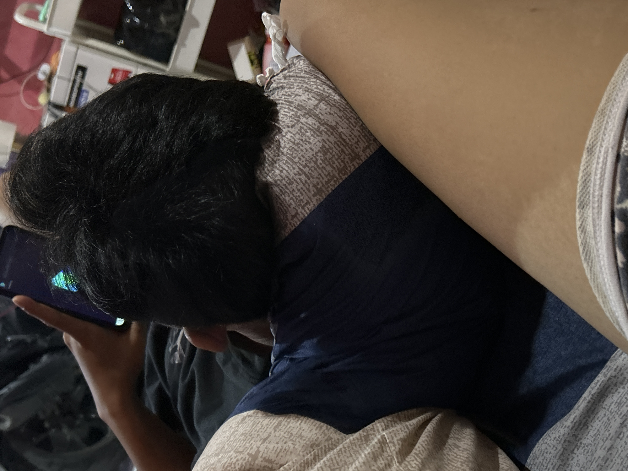 The image shows a close-up of a person's head and shoulder as they lie down and play a game on their phone. The person has dark, short hair and is wearing a dark blue t-shirt. They are holding a smartphone in their left hand, with their thumb on the screen. The phone screen displays a colorful game with blue, green, and purple hues.
The person is lying on a bed or sofa, with a patterned pillow supporting their head. The pillow has a light brown and white abstract pattern. To the right of the person, a part of their leg is visible, with light-colored skin. They appear to be relaxed or engrossed in the game. The background is blurred and shows some shelves with items, suggesting an indoor setting, possibly a bedroom or living room. The lighting suggests it might be nighttime or indoors with artificial light.
There is no visible text in the image. The overall mood is casual and focused on the activity of playing a mobile game. The setting appears to be a home environment in Depok, Indonesia.