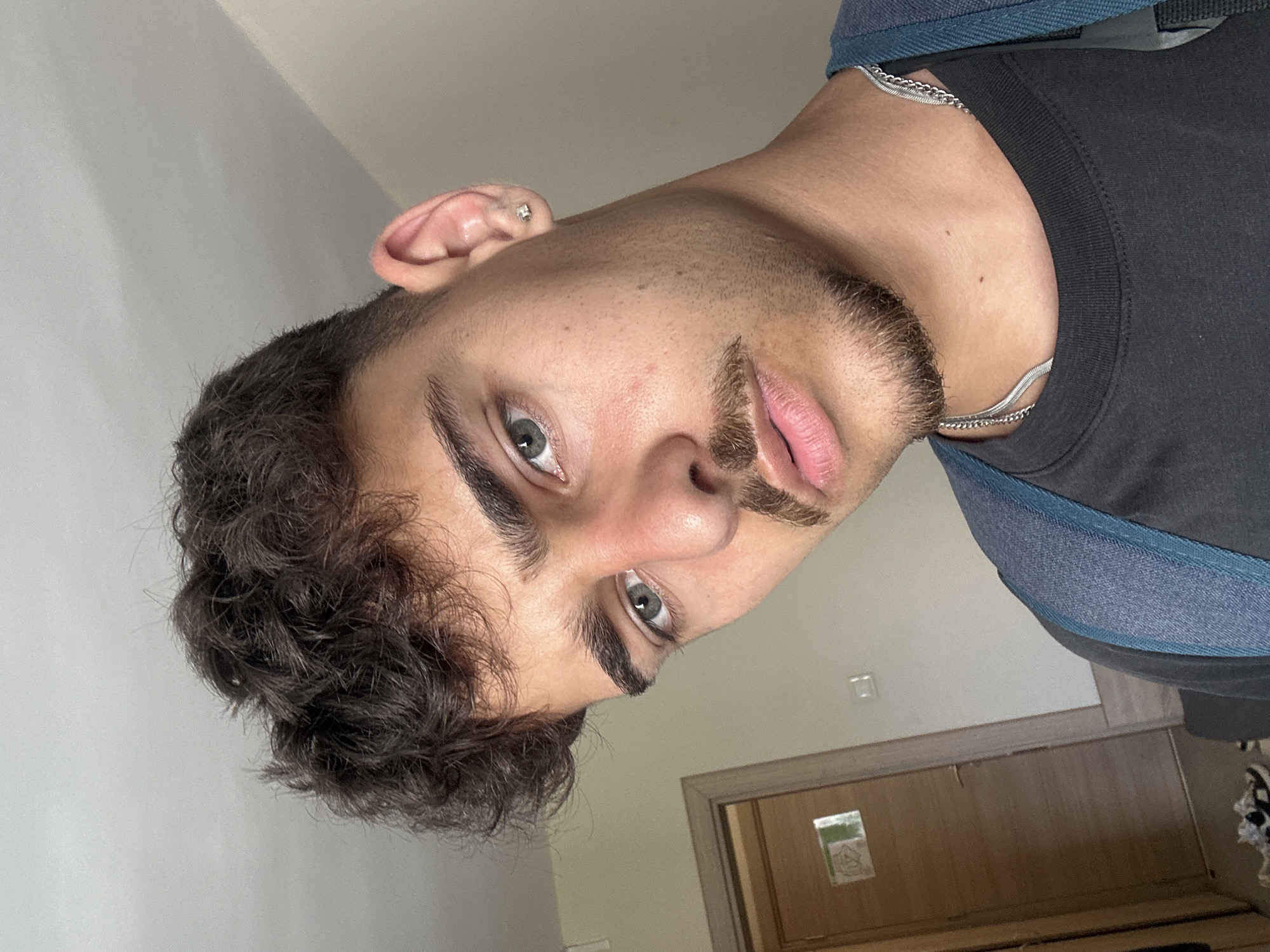 A young adult male with dark curly hair, facial hair (mustache and goatee), and light-colored eyes (possibly green or hazel) is the primary subject of the image. He is wearing a dark t-shirt and a blue backpack. A silver chain necklace is visible around his neck. He has a piercing in his left ear. The background shows a light-colored wall, a doorframe, and a partially visible door with a sign on it. Given the style of the door, sign and wall paint the location could possibly be a hotel room. There is no definite evidence of this being taken in Sofia, Bulgaria.