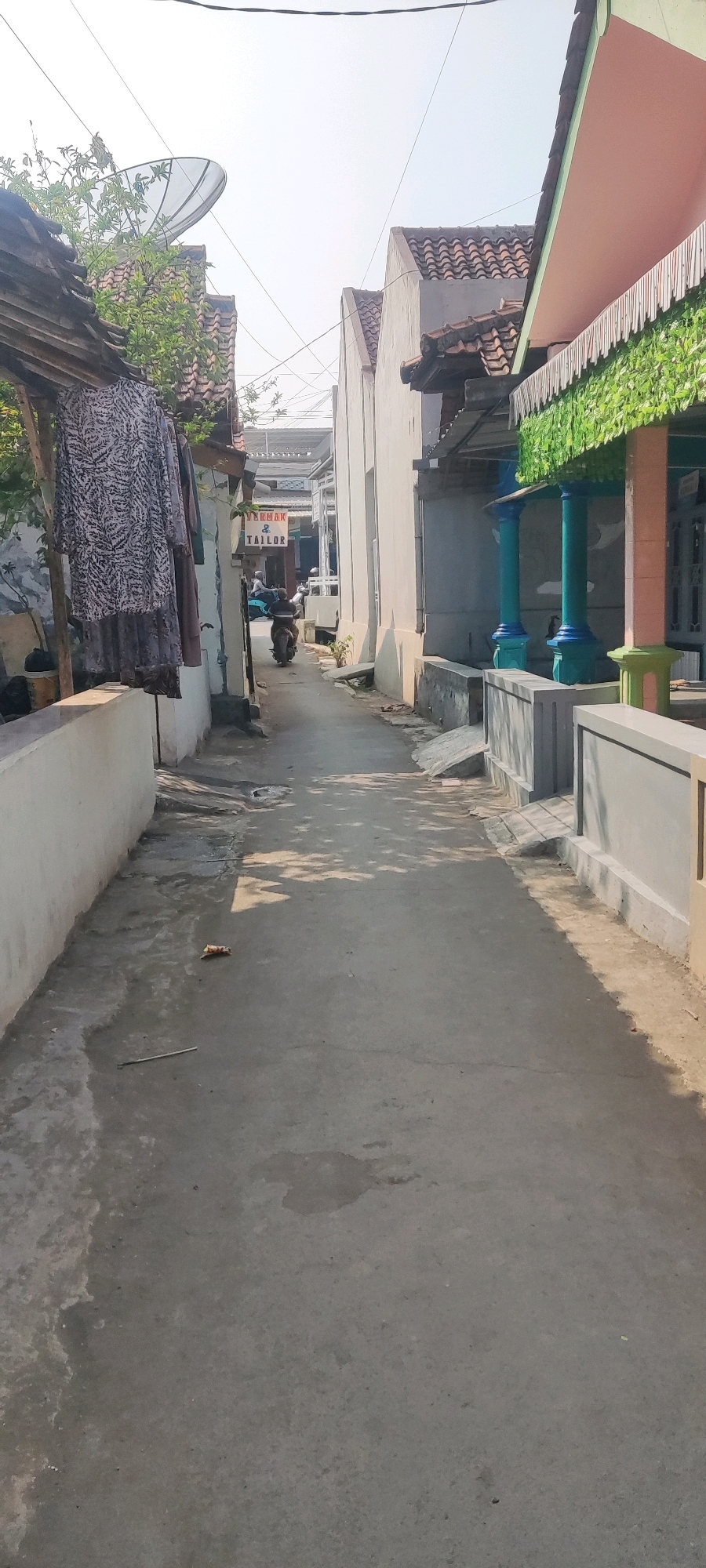 The image depicts a narrow, paved alleyway in the city of Gebang, Indonesia. The alley is flanked by residential buildings with tiled roofs. On the left, a white wall borders the alley, with a wooden structure supporting a clothesline holding several garments. A satellite dish is visible on the roof of a building to the left, above some greenery. A sign reading "TYREMAH & TAILOR" is mounted on a building further down the alley on the left. On the right, a building features a decorative facade with green foliage and blue pillars. The alleyway is illuminated by sunlight, casting shadows on the ground. A person is riding a scooter down the alley, moving away from the viewer. A small piece of litter, possibly a food wrapper, lies on the ground in the middle of the alley. The environment appears to be a residential or mixed-use area within a town or city.