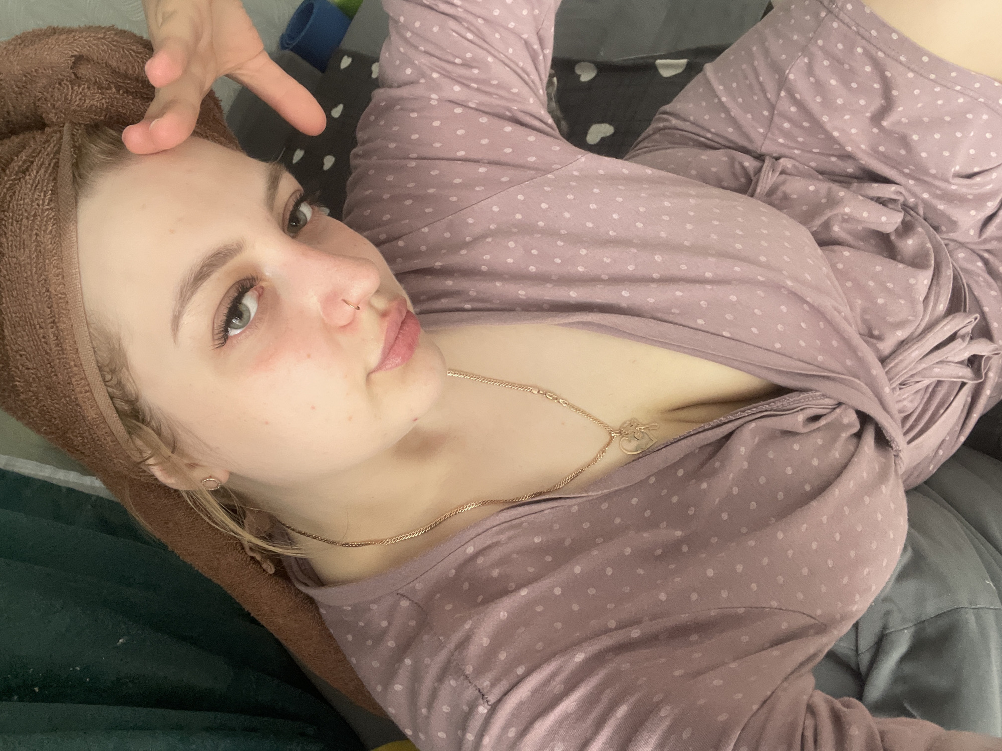 A fair-skinned female with a brown towel wrapped around her head is lying on a bed, looking at the camera. She is wearing a long-sleeved, mauve-colored pajama top with white polka dots and a gold necklace. Her left hand is raised towards her forehead. The background includes a gray pillow, a pillow with black hearts on a white background, and a blue object. The location cannot be determined from the image.