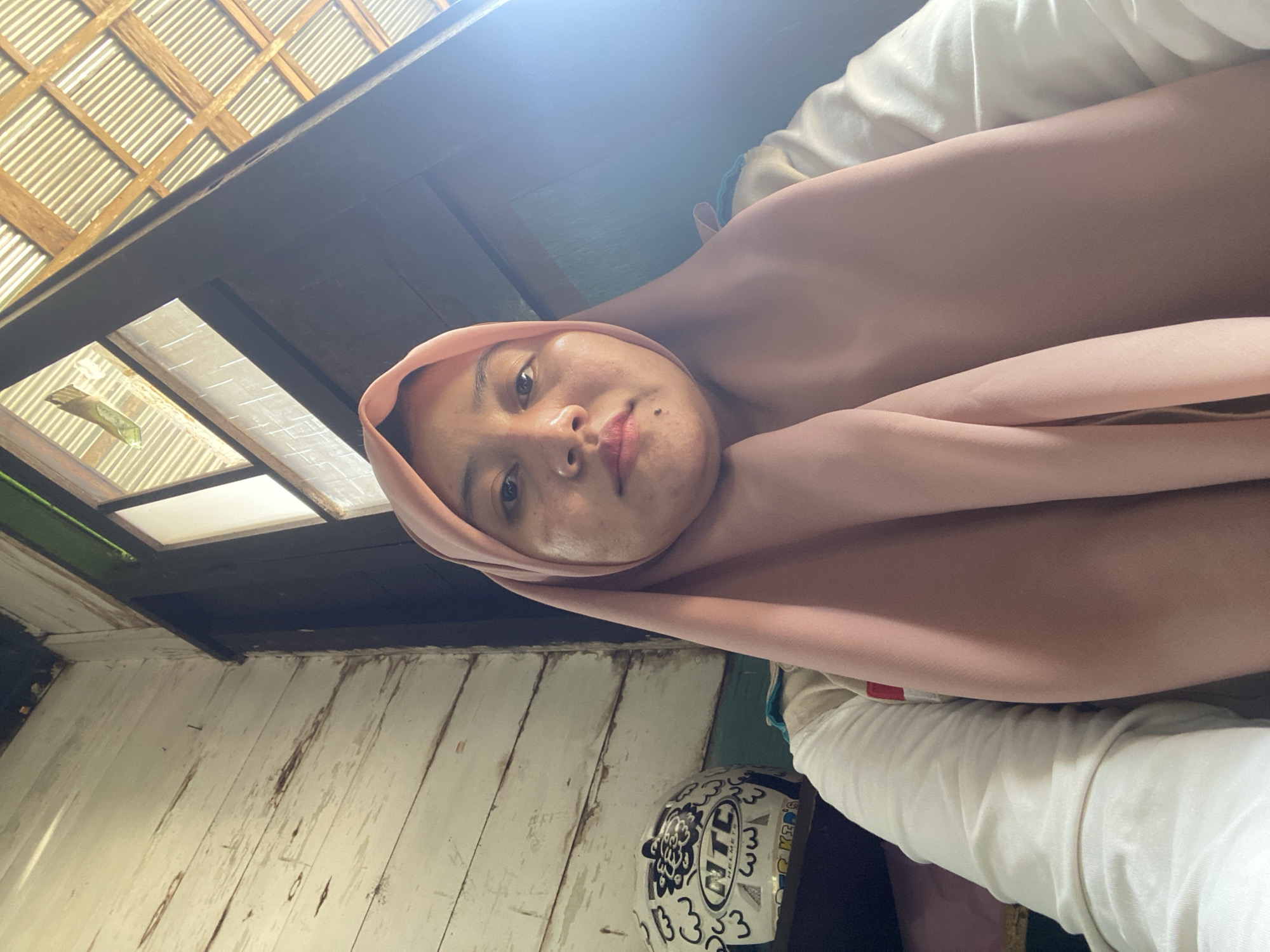 A young woman wearing a pink hijab and a white long-sleeved shirt is taking a selfie in a rustic, interior setting. The wall behind her is made of weathered, white-painted wooden planks, showing signs of age and wear. To her left, a white motorcycle helmet adorned with black and blue drawings and logos such as "NTC HELMETS" is visible. The background also includes a dark wooden door or window frame and a corrugated metal ceiling, suggesting a simple or rural dwelling. The lighting indicates it is daytime, with sunlight streaming in from the right, creating a bright, slightly overexposed area. The woman's expression is neutral, and her face shows some skin blemishes.
