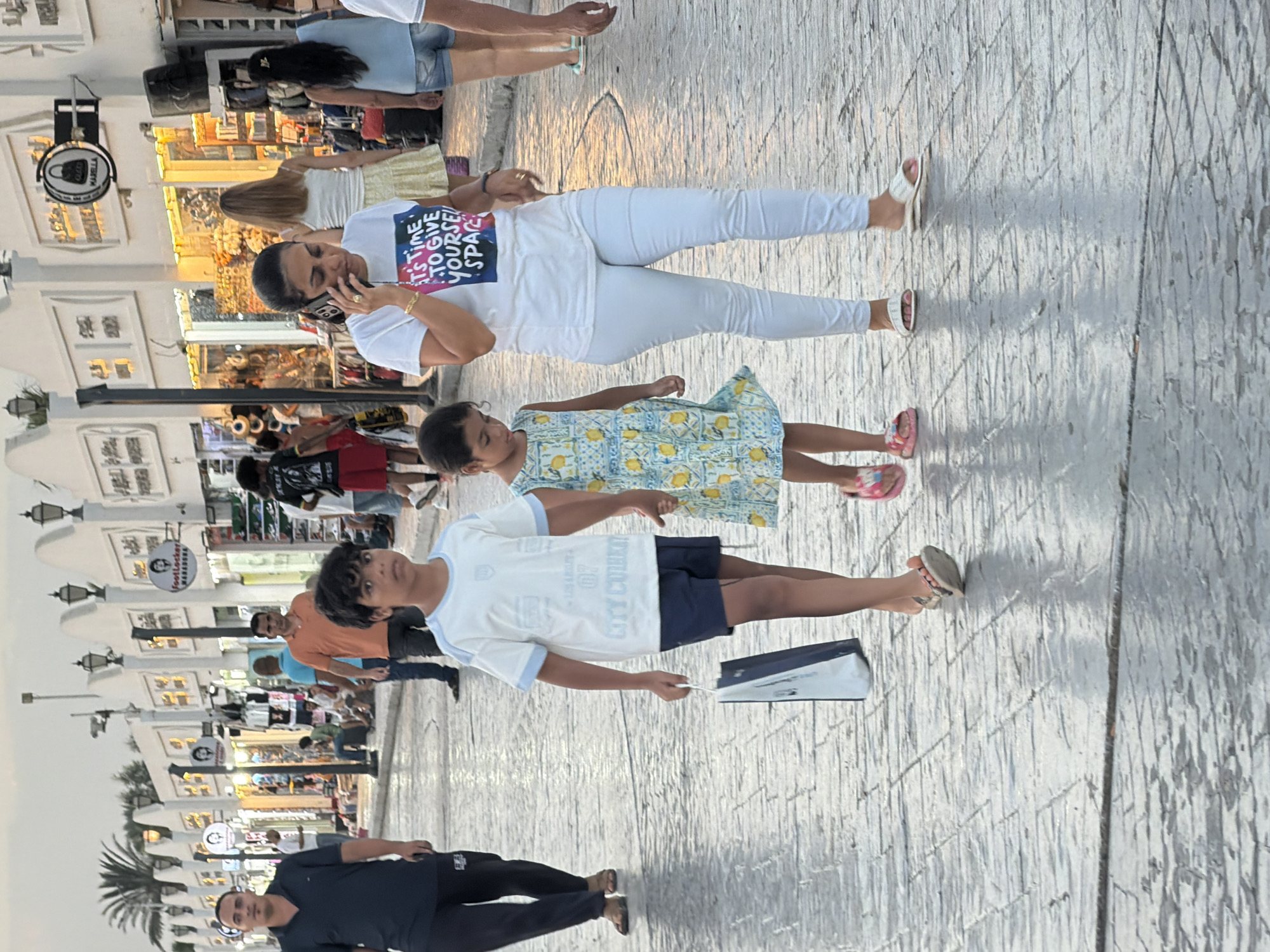 In Sharm El Sheikh, Egypt, a diverse group of people walk along a patterned cobblestone street lined with shops. In the foreground, a young boy in a white t-shirt and dark shorts walks left, holding a shopping bag. Beside him, a young girl in a yellow and blue patterned dress walks right. Behind them, a woman in a white t-shirt with colorful text and white pants talks on her phone, wearing white sandals. Further in the background, other individuals are seen walking and standing, including a man in a dark polo shirt and dark pants, and a man in an orange t-shirt. Shops with illuminated displays and signs are visible along the street. The sky is overcast, suggesting it is likely late afternoon or early evening.