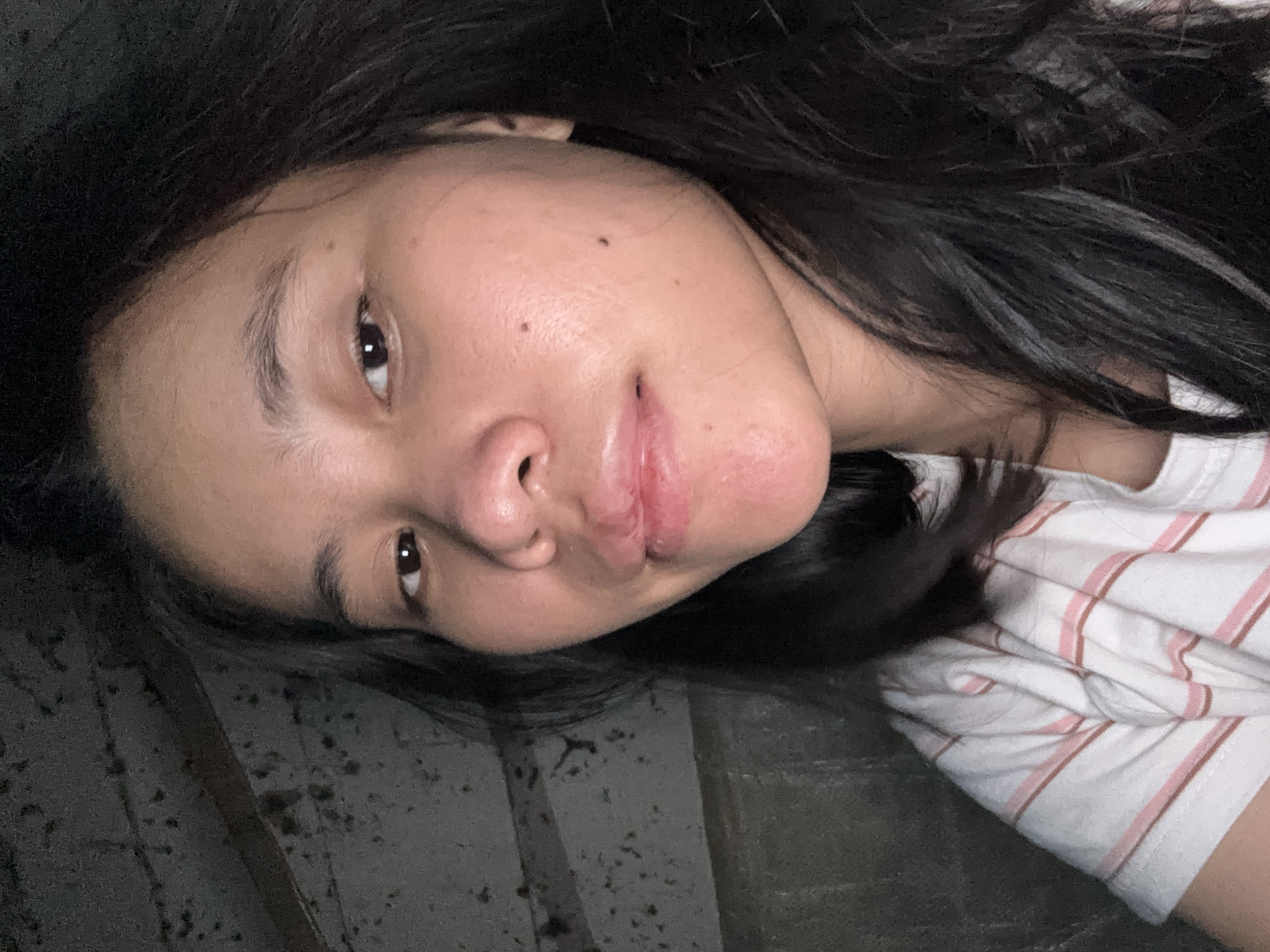 A close-up, slightly high-angle, medium-format selfie shows a young woman from the chest up. She has long, dark, straight hair that falls over her shoulders and frames her face. She is wearing a white t-shirt with thin pink and brown stripes. Her skin is fair with some freckles visible on her right cheek. Her lips are slightly parted and appear dry. The background is dark and appears to be a textured, possibly concrete wall with some horizontal lines, hinting at an indoor, possibly unfinished, setting. There are no other people or objects of note in the frame. The lighting is dim, casting soft shadows on her face. No specific location within East Jakarta, Indonesia can be determined from the visual information provided.