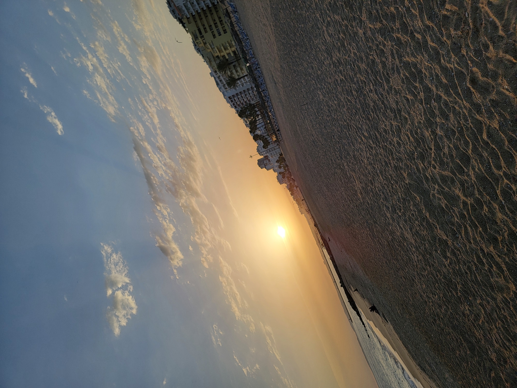 This image depicts a sunset scene on Quarteira beach in Portugal. The sun, a bright orb, is low in the sky, casting a warm, orange glow across the scene and creating a gradient of color in the sky from bright yellow near the sun to blue at the top. Wispy clouds are scattered across the sky, some catching the golden light of the sunset. The beach sand, in the foreground, shows the texture of small waves and ripples.
In the middle ground, the gentle waves of the Atlantic Ocean meet the shore. A dark, rocky breakwater extends into the sea from the left side of the image. Along the coastline in the background, there are several multi-story buildings, likely hotels or apartments, characteristic of a coastal resort town. Palm trees and beach umbrellas are visible in front of these buildings, suggesting a developed tourist area. A few distant figures can be seen on the beach and near the water's edge. The overall impression is one of a peaceful evening at a popular European beach destination.
