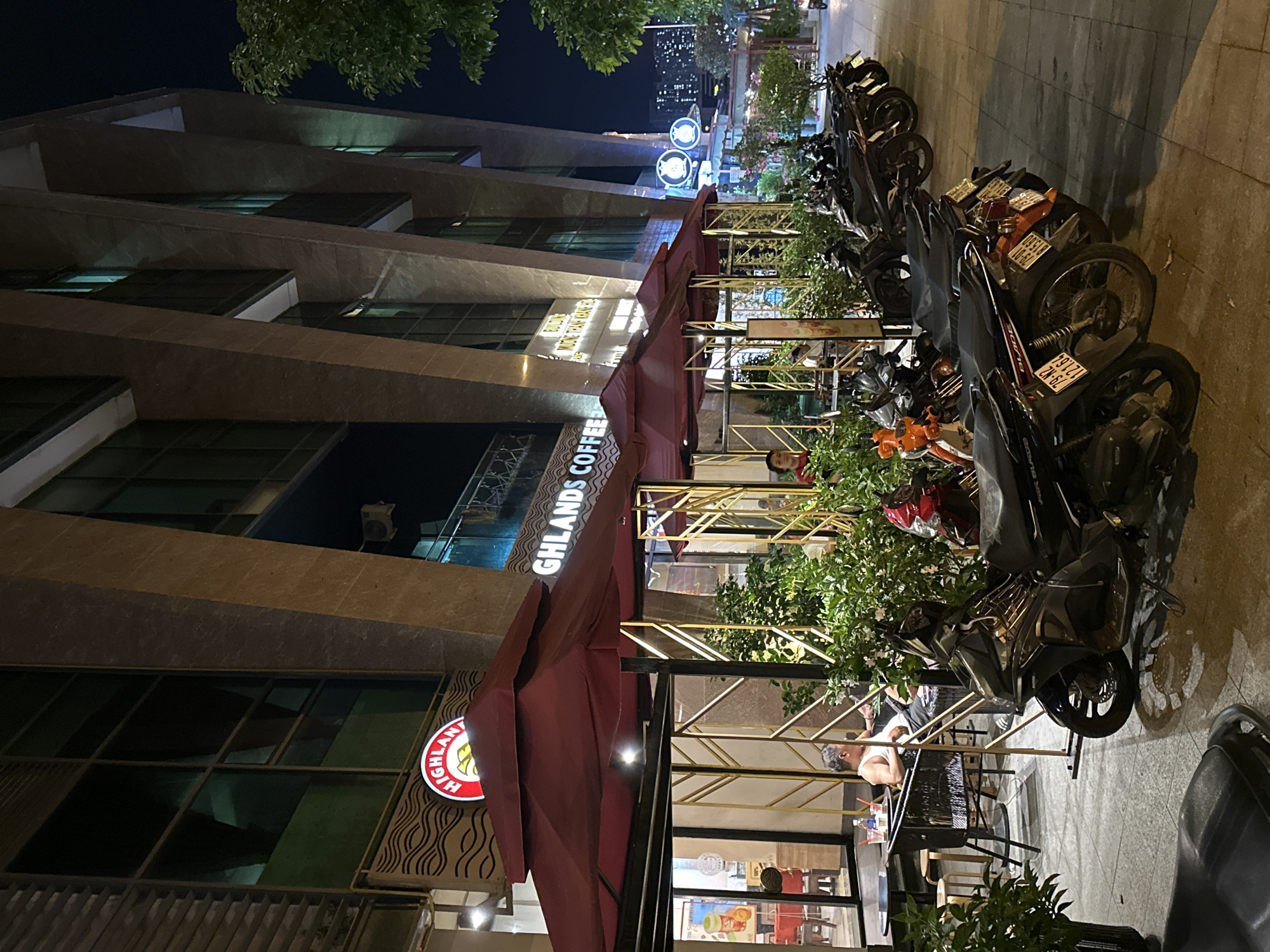 The image captures an outdoor seating area of a Highlands Coffee shop at night in Nha Trang, Vietnam. The establishment is situated in front of a modern building with angled architectural elements. A prominent red awning, supported by golden metal frames, covers the seating area. Several motorcycles are parked on the sidewalk in front of the coffee shop. Two individuals are visible seated at a table under the awning, and another person stands near the entrance. The lighting suggests it is nighttime, with artificial lights illuminating the scene. The overall impression is a typical urban scene in Vietnam, with a focus on a popular coffee chain and its surrounding environment.