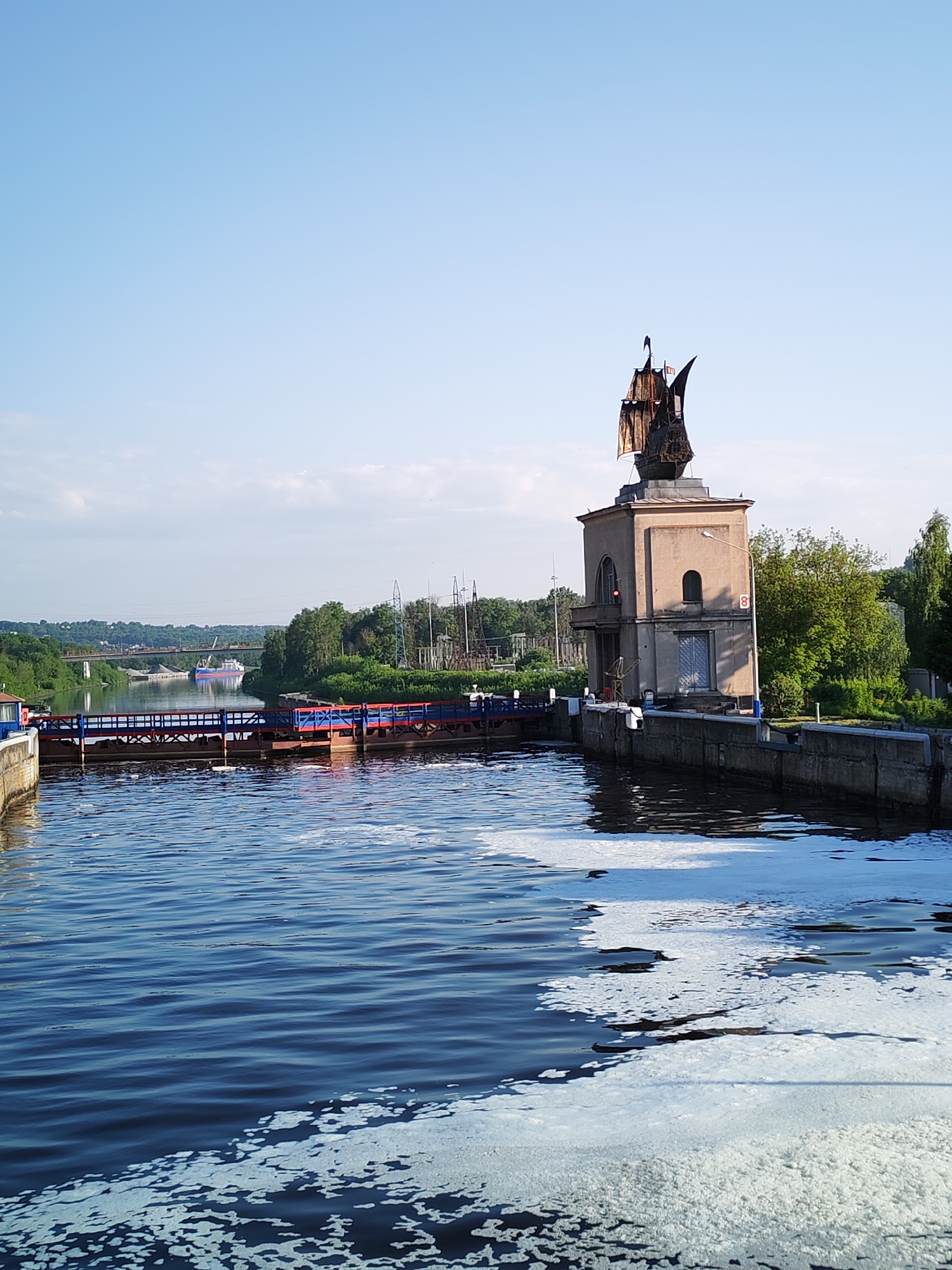 The image depicts a river scene in Yakhroma, Russia. A statue of a ship atop a building stands on the right bank. The river is actively flowing, with white foam evident in the water. A lock system is visible, painted red and blue, positioned between the viewer and the far bank. Across the river, trees line the bank. In the distance, a bridge spans the river. The sky is clear and bright blue, suggesting a sunny day.