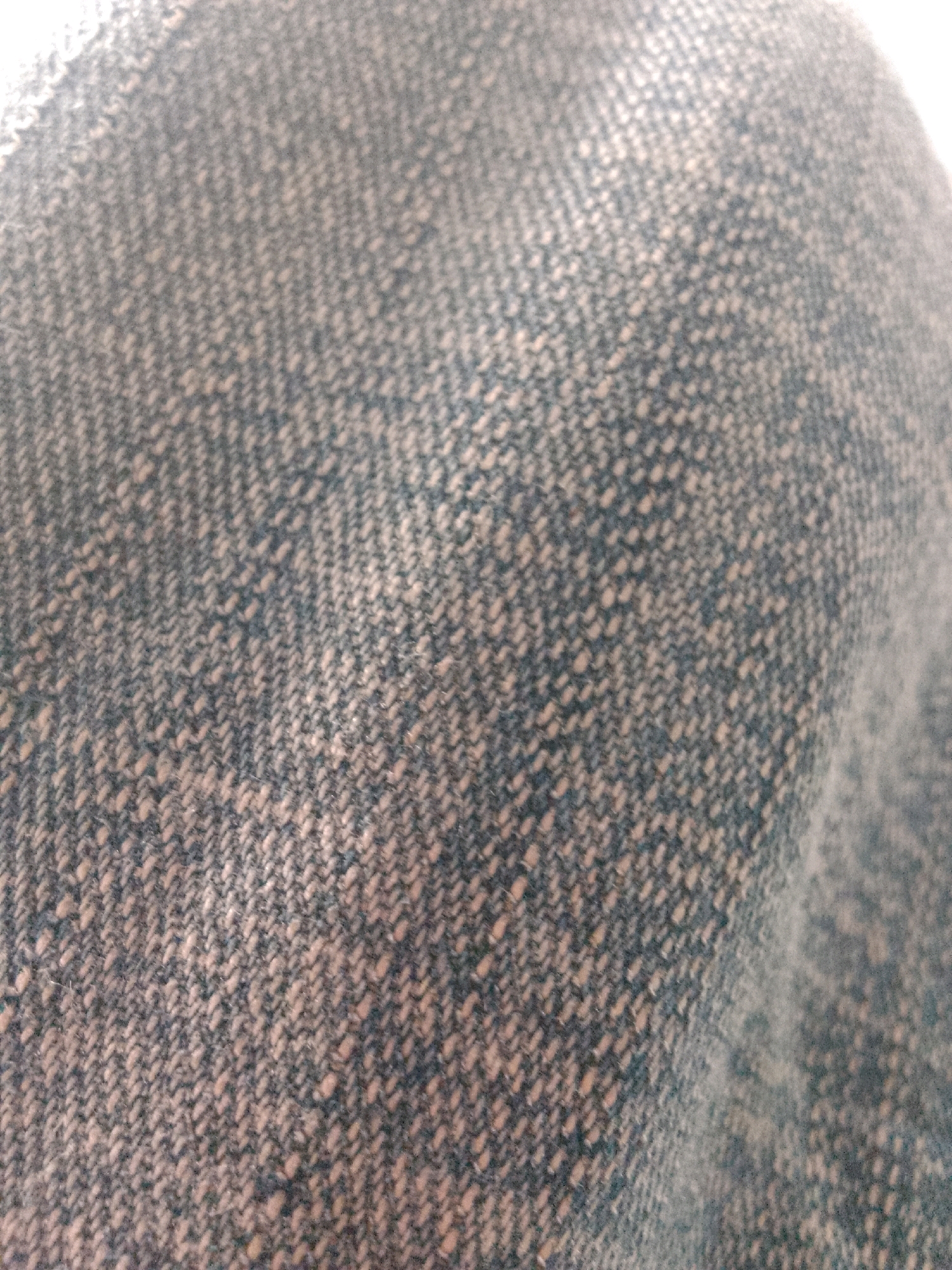 The image displays a close-up, abstract view of a textured fabric. The fabric appears to be woven with a mix of dark blue and light beige or off-white threads, creating a speckled or tweed-like pattern. The weave is visible in a diagonal herringbone or twill pattern. The lighting suggests a soft, diffused source, possibly from the upper right, causing some areas to be brighter and others to be in shadow, emphasizing the texture. There are no people, objects, actions, or interactions discernible in the image, as it focuses solely on the surface of the material. The location in Trenggalek, Indonesia, cannot be determined from the visual information provided.