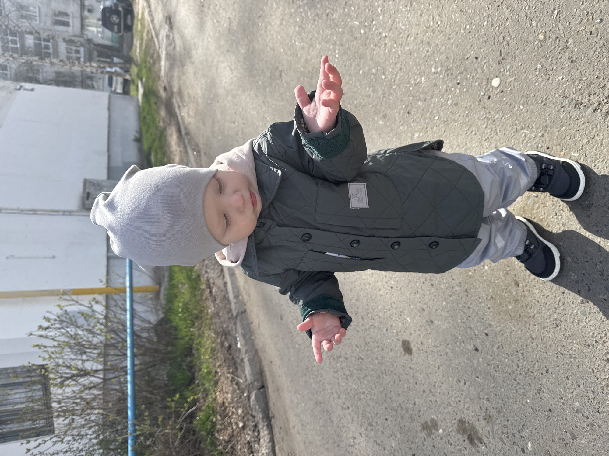 A toddler in a grey knit hat, khaki quilted jacket, light grey pants, and black sneakers stands on a concrete sidewalk outdoors. The child's hands are raised. Behind the child are buildings and some sparse foliage. The location is Chaikovsky, Russia.