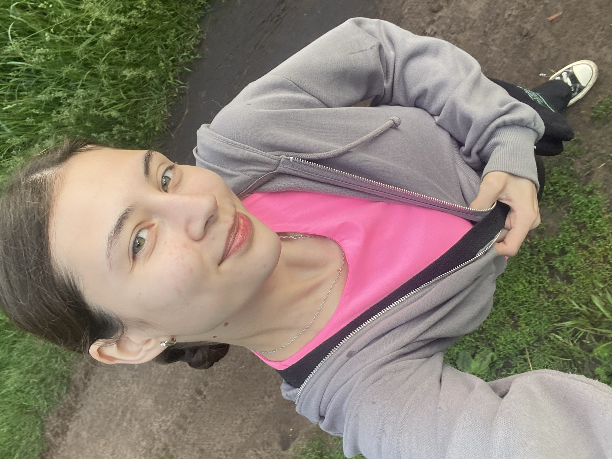 A young Caucasian woman with dark hair pulled back in a braid is taking a selfie outdoors. She is wearing a pink t-shirt under a grey zippered hoodie and black pants with a green design. Her hair is styled in a braid that is visible over her left shoulder. She has a silver necklace on and a small hoop earring in her left ear. Her skin is fair and she has a light smile.
The background consists of a dirt path and green grass. The lighting suggests it is daytime, possibly late afternoon or early evening, with soft natural light.
It is not possible to determine the specific location within Votkinsk, Russia, from the provided image alone. The scene depicts a natural or semi-natural outdoor environment, consistent with a park, trail, or rural area.