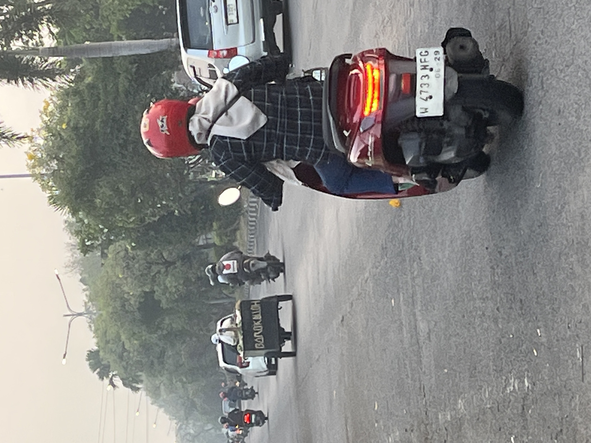 The image captures a street scene in Kludan, Indonesia, likely during the daytime given the ambient light. The foreground features a person riding a red scooter, wearing a red helmet and a checkered long-sleeved shirt. The scooter has a visible license plate that reads "W 4733 HFG 06-29". In the background, several other motorbikes and a white car are visible, navigating the road. One motorbike is pulling a small cart with "BAROKALLOH" written on its side. The environment appears to be a road with trees and streetlights lining the sides, suggesting an urban or suburban setting. The overall atmosphere is hazy, possibly due to pollution or the time of day.