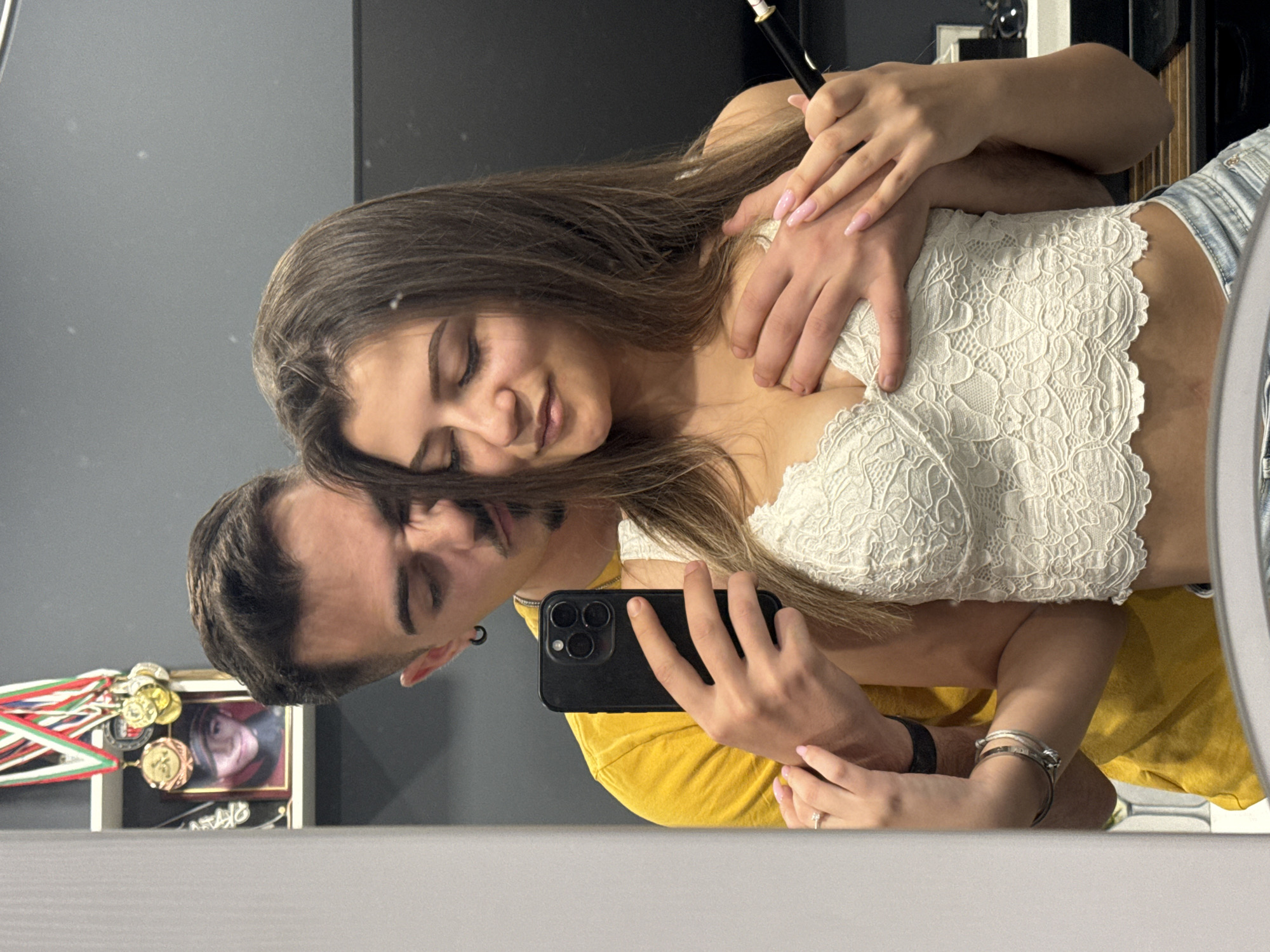 A couple is reflected in a mirror, possibly in a private residence. The woman has long, brown hair and is wearing a white lace top and light-wash denim shorts. The man, with short dark hair and a mustache, is wearing a yellow t-shirt and holding a black smartphone. The woman is holding a cosmetic tool. Behind the couple, a shelf holds a framed portrait and a display of medals with ribbons. The location is likely within Sofia, Bulgaria.