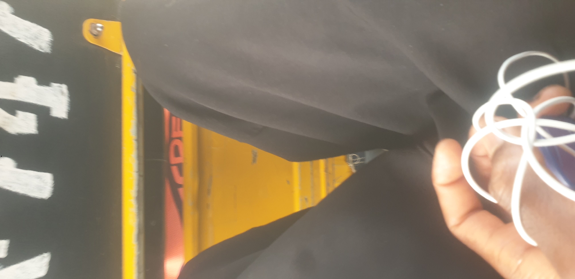 The image shows a close-up view of a person's lap wearing dark pants, with their hand holding white earbud wires. In the background, a yellow surface with a black stripe and white lettering is visible. Partially obscured red text on the yellow surface reads "CRE". A yellow metal bracket with a silver screw is attached to the black stripe. The scene appears to be inside a vehicle, possibly a tuk-tuk or a similar mode of transport. The person's hand and the earbuds suggest they may be listening to music or a podcast. The limited visibility of the environment makes it difficult to pinpoint an exact location within Barakin Lerawi, Nigeria, but the presence of distinctive vehicle elements suggests it is a common mode of transport in the area.