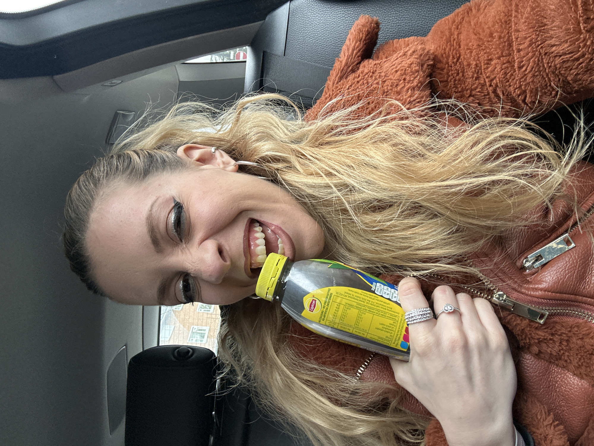 The image shows a woman in a car, holding a bottle of Lipton iced tea. She is smiling and looking at the camera. She is wearing a brown, fur-lined jacket and has blonde hair. She is wearing earrings and rings on her right hand. The background of the image shows the interior of the car and a glimpse of buildings outside, but without further context the exact location in the city of London, United Kingdom cannot be determined.