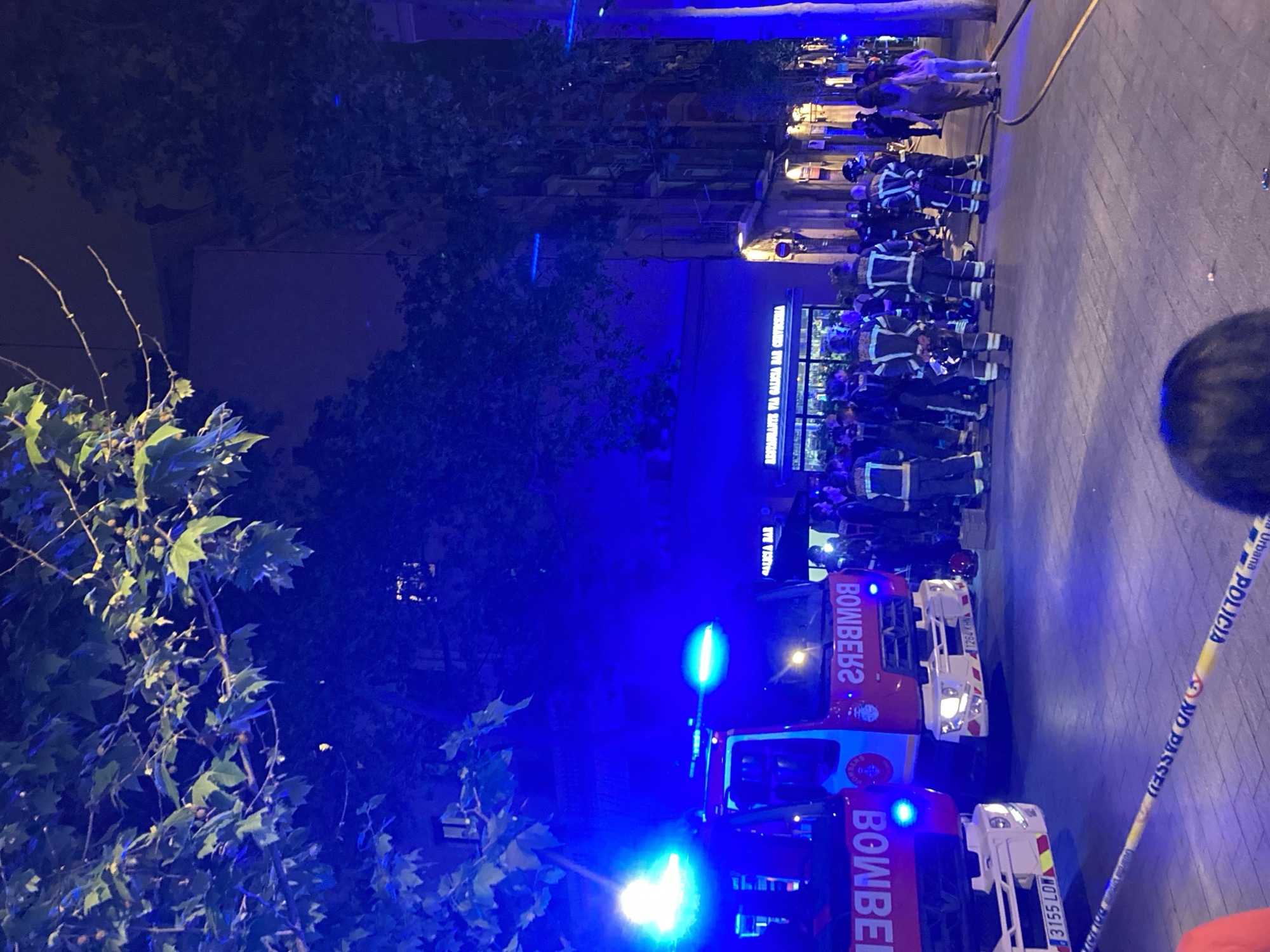 At night, in an urban setting, the scene is illuminated by bright blue emergency lights. Two fire trucks labeled "BOMBEROS" are parked with their lights flashing, indicating an ongoing situation. A group of firefighters in uniform gathers near the front of a building, possibly a restaurant or bar, identified by a sign that reads "RESTAURANTE VIA GALICIA BAR". Police tape labeled "POLICIA NO PASSED" cordons off a section of the area, suggesting a restricted zone. Trees with green leaves are visible to the left of the scene, adding a natural element to the urban environment. A few people stand along a sidewalk.