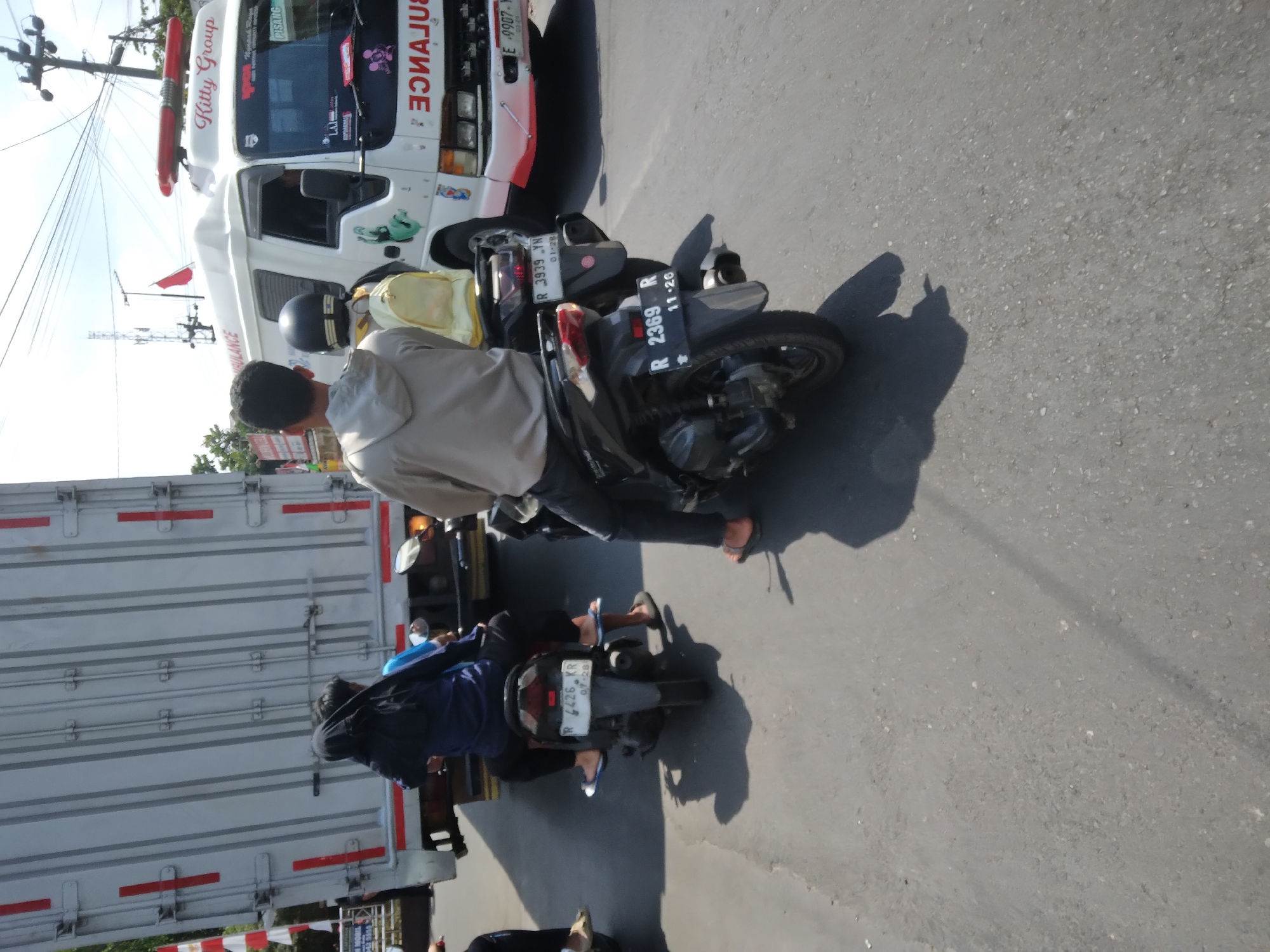 The image captures a street scene in Desa Pamijen, Kecamatan Sokaraja, Indonesia, on a sunny day. The foreground features two motorcycles. The one on the left is ridden by a person wearing a blue top and black pants, with a black hijab. The motorcycle on the right is occupied by a person in a grey hoodie and black pants, with a person in a yellow jacket and black helmet on the back. Both motorcycles have visible license plates. The license plate on the left motorcycle reads "R 4426 KR 07-26". The license plate on the right motorcycle reads "R 2369 R 11-26". In the background, a large white truck dominates the scene, with a white ambulance with red lights on top to its right. The ambulance has "Kitty Group" written on its side. The road surface is asphalt, and shadows are cast by the vehicles and people, indicating the time of day. There are some overhead wires and utility poles visible in the background.