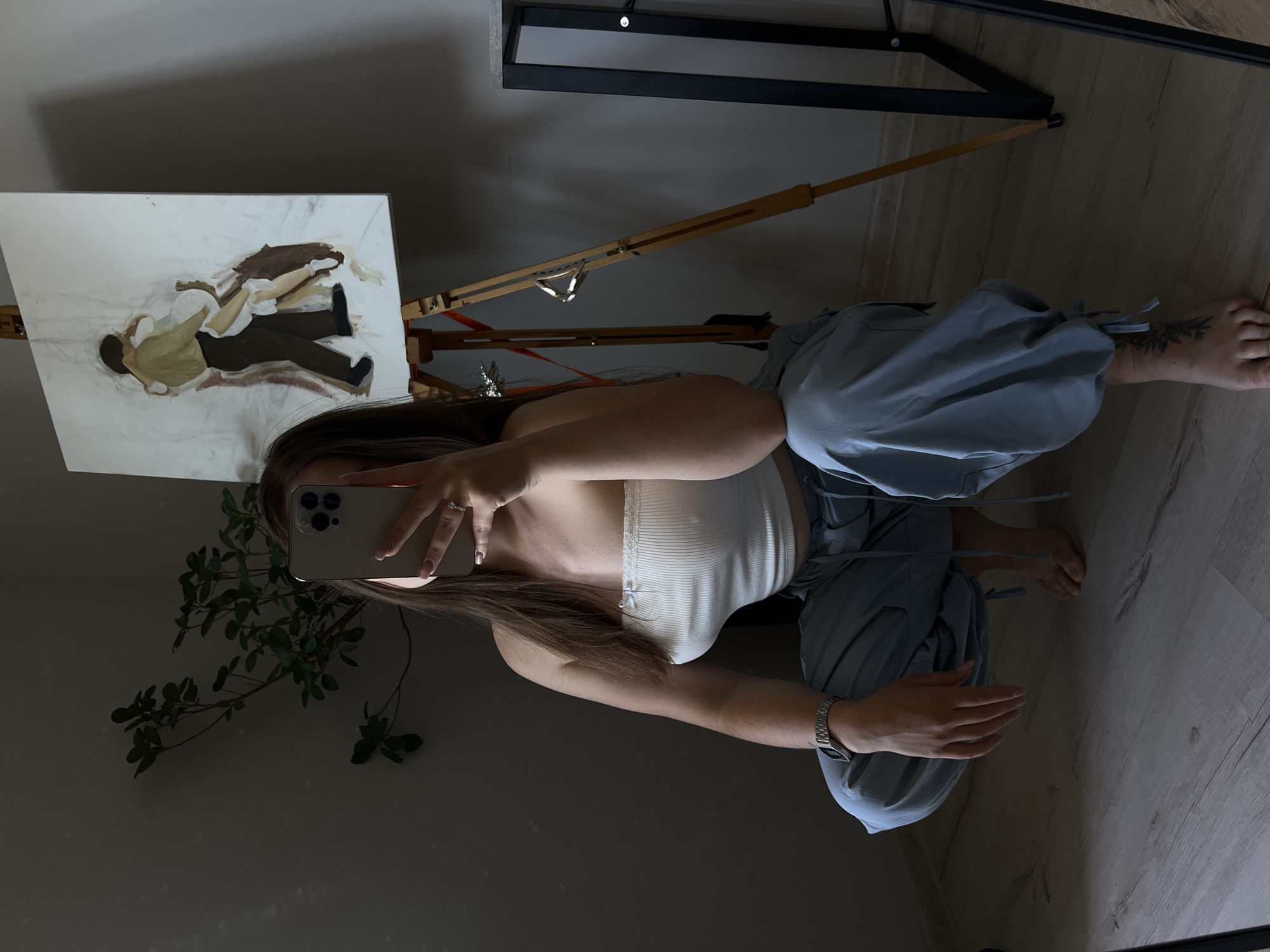 A young woman with long brown hair is taking a selfie in a room. She is wearing a white tube top and light blue cargo pants. She is squatting on a wooden floor in front of a painting on an easel. The painting depicts two figures dancing. To her left is a potted plant. To her right, a part of a black and silver frame is visible. The lighting in the room is dim, with a strong shadow cast from the easel and painting onto the wall. There are no discernible signs of the city of Ternopil, Ukraine.
