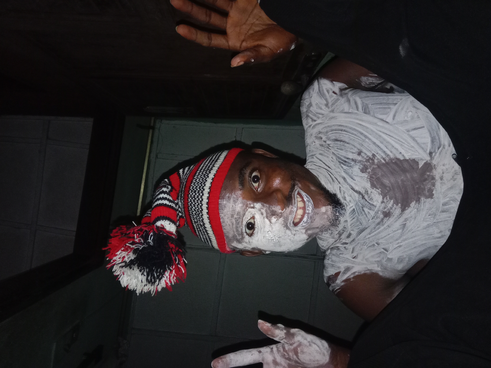 A man is seen from a low angle, partially covered in white paint on his face and body. He is wearing a knitted cap with red, black, and white stripes and a pom-pom. His hands are raised, also partially covered in paint. The environment appears to be indoors with dark surroundings. The location is Unknown, Unknown.