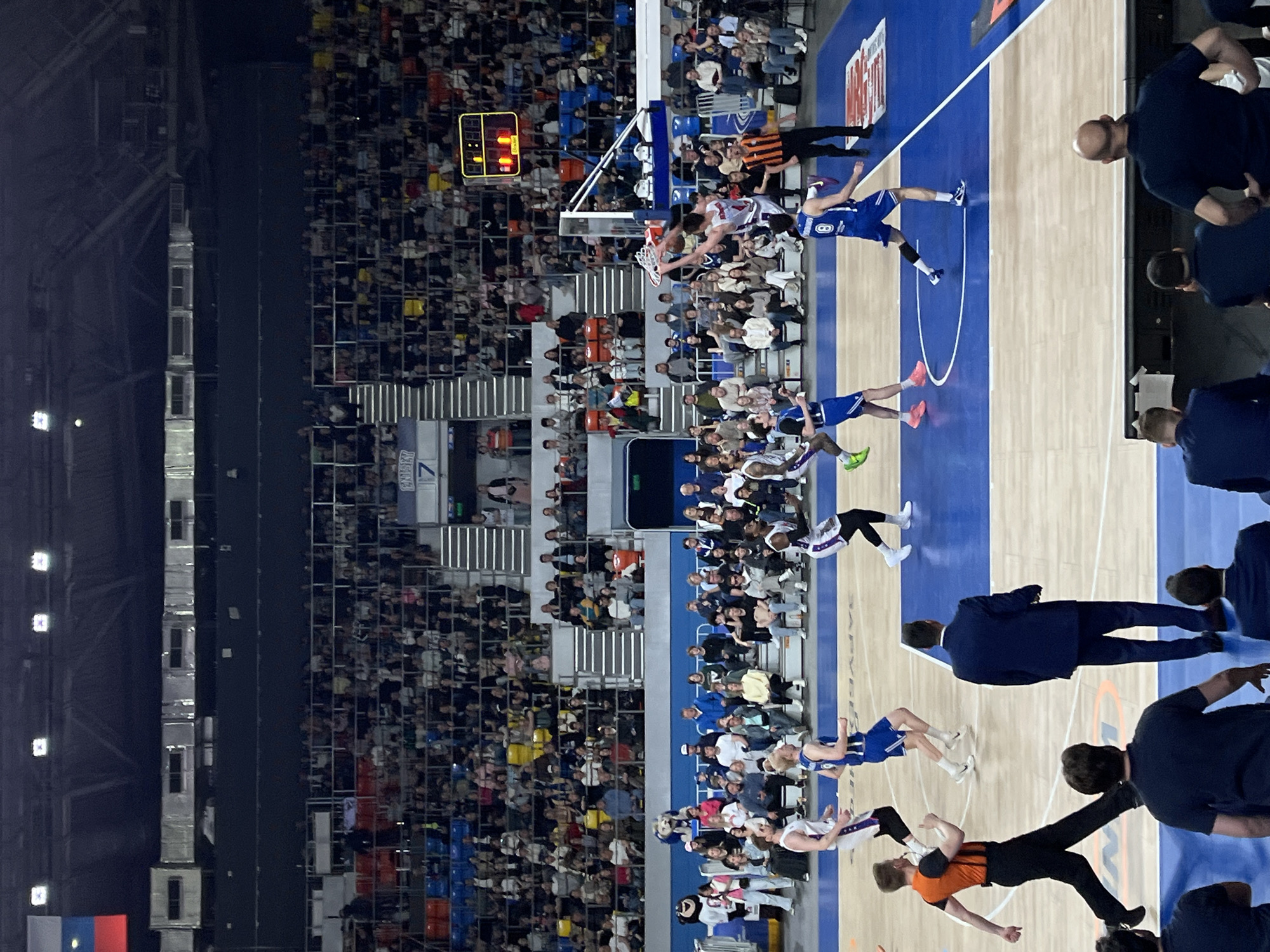 The image depicts a basketball game in progress within a large arena in Krasnoyarsk, Russia. Players in blue and white uniforms are actively engaged in gameplay on a polished, marked basketball court with blue borders. One player is shown attempting to score at the basket near the scoreboard. An official in an orange and black striped uniform observes the play closely. Surrounding the court are seated spectators filling the arena, with rows of people on multiple tiers. The arena is well-lit with overhead lighting fixtures.