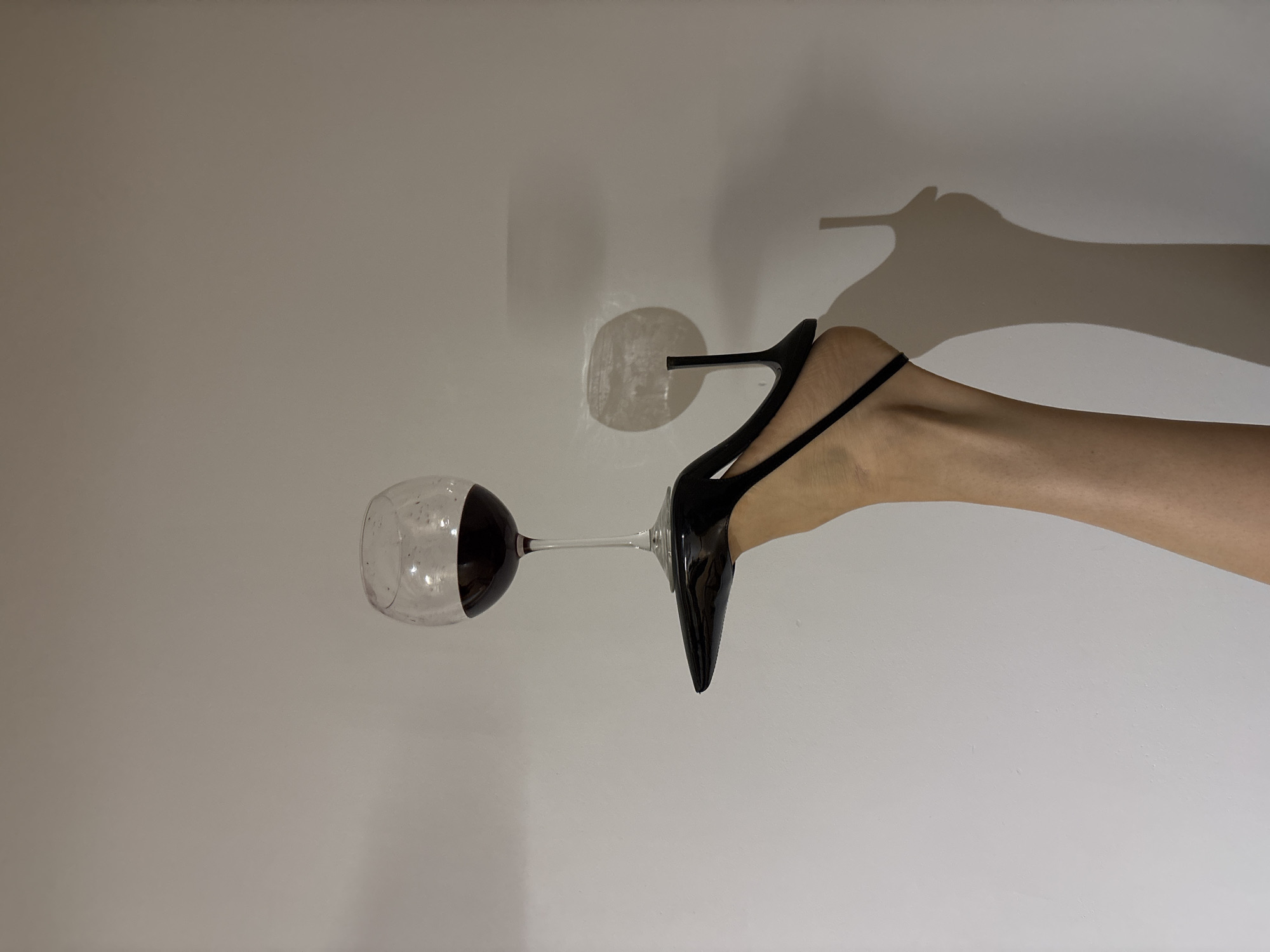 The image displays a stylized, rotated composition featuring a bare human leg and foot wearing a black high-heeled shoe, delicately balancing a wine glass filled with red wine. The scene is set against a plain, light-colored wall, which provides a minimalist background.
The foot, clad in a sleek black stiletto with a thin strap across the instep, is positioned horizontally across the mid-right of the frame. The slender stem of the wine glass is either held or balanced by the shoe's heel or the foot itself, extending to the left. The glass contains a notable amount of dark red liquid, strongly suggesting wine.
Prominent shadows are cast on the light wall to the right, forming a large, elongated silhouette of the leg and shoe, adding depth and an abstract element to the composition. A distinct circular reflection or light anomaly is also visible on the wall behind the shoe's heel. The entire image appears to be rotated 90 degrees counter-clockwise, as the leg and wine glass are oriented horizontally.
The setting is an indoor space, indicated by the plain wall and shadow patterns. No other objects, people, or specific location cues are visible. There is no discernible text anywhere in the image. The time of day cannot be determined, but the lighting suggests an intentional setup. The overall mood is artistic and focused on form and balance.
