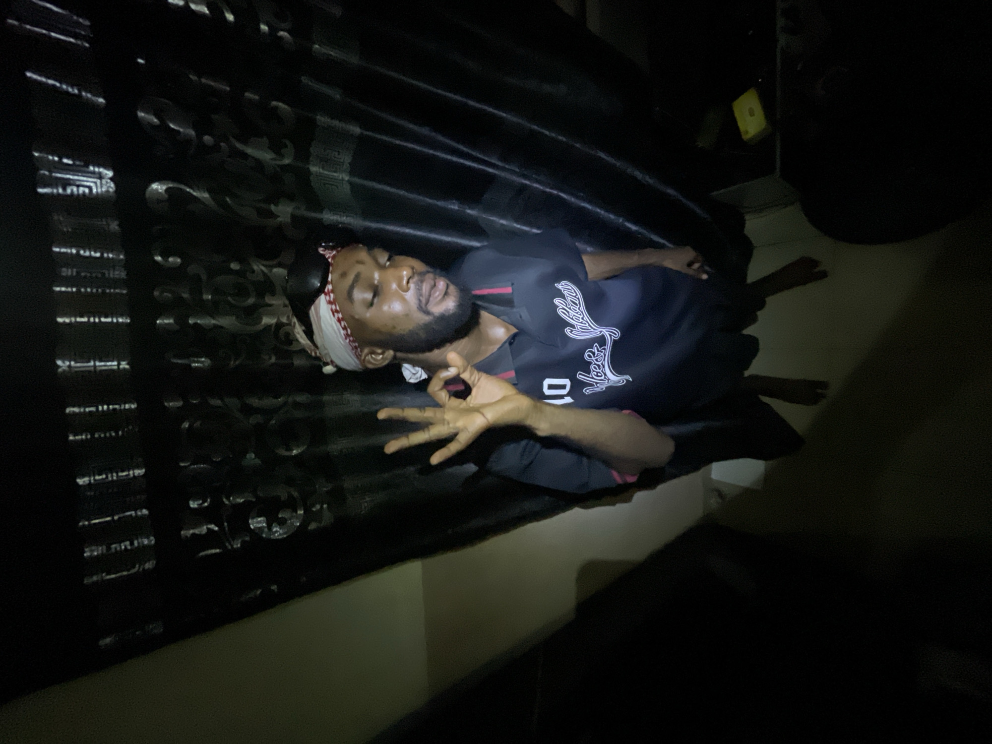 A man is lying on a surface, possibly a bed, against a black background. He is wearing a dark shirt with white writing, a white and red headband, and has a beard. His eyes are closed. He is making a hand gesture with his right hand. The lighting is dim. The location is Lokoja, Nigeria is not discernible from the image.