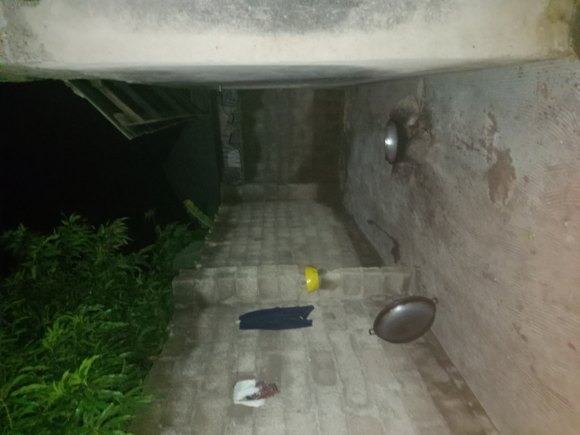 The image depicts a dimly lit outdoor courtyard or alleyway, likely in Jalingo, Nigeria. A rough, cinder block wall runs along the left side of the frame, adorned with two pieces of laundry hanging on it: a patterned cloth on the left and a dark blue garment to its right. To the right of the dark blue garment, a yellow bowl is attached to the wall. In the foreground, a large, dark, circular pot or pan with handles rests on the ground, leaning against the wall. Further into the courtyard, a smaller, metallic bowl is set on stones, possibly part of a makeshift stove, with visible remnants of ash or soot beneath it. The ground is paved with concrete, showing some damp patches and discoloration. Lush green foliage overhangs the wall on the left side of the image. To the right, a light-colored wall with a rough texture forms the boundary of the courtyard. A sloped, corrugated roof structure is visible in the upper right portion of the image. The overall impression is one of a functional, albeit basic, outdoor space, possibly used for cooking or domestic activities.