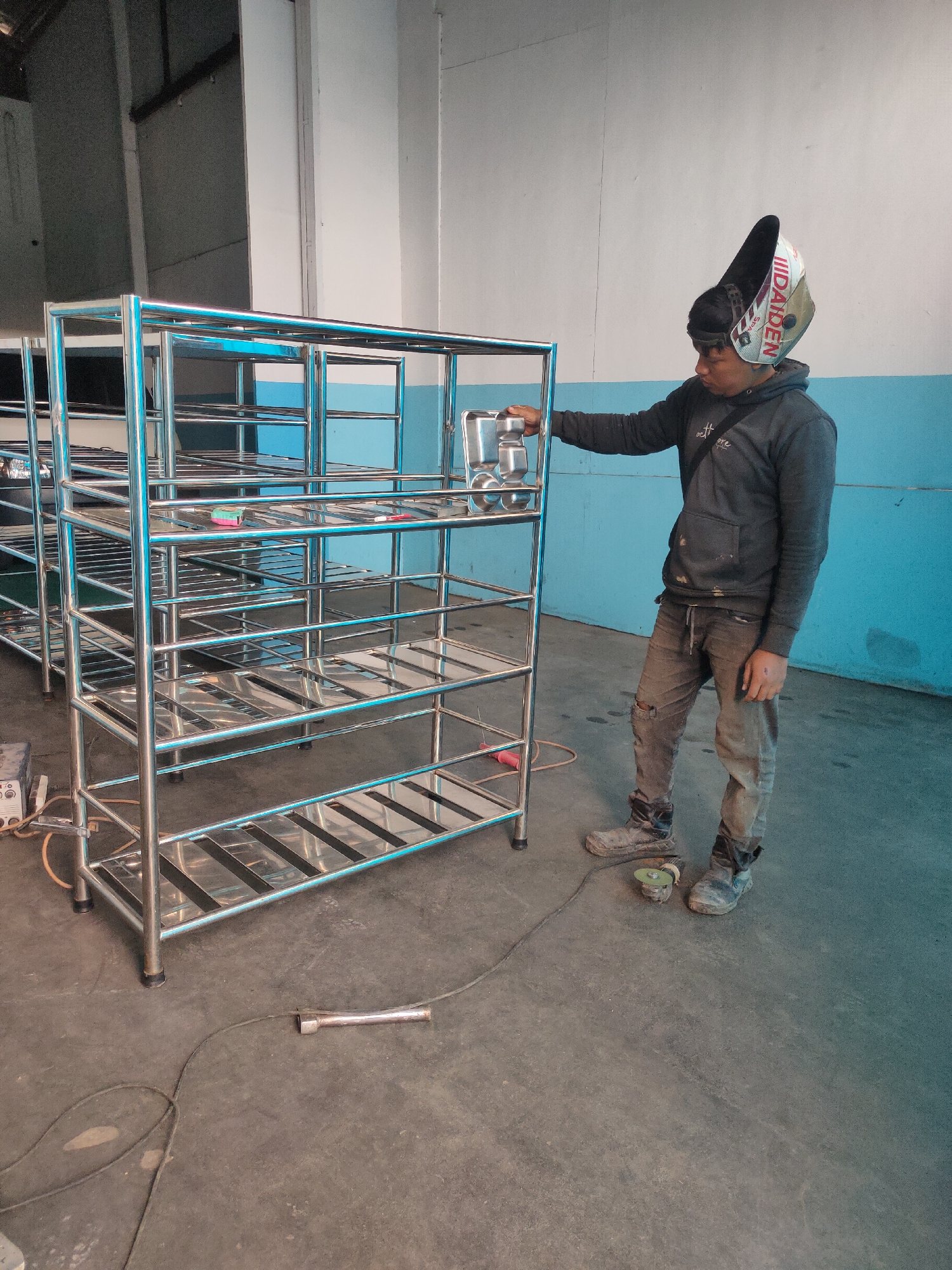 A man, wearing a welding mask tilted upwards on his head, stands in a workshop in Sumedang, Indonesia. He is wearing a dark hooded sweatshirt and worn-out pants, with muddy boots on his feet. The man is leaning on a tall, multi-tiered stainless steel shelving unit. Several similar shelving units are visible behind him. A welding machine and electrical cords are on the floor near the shelves. The walls of the workshop are painted light gray and blue.