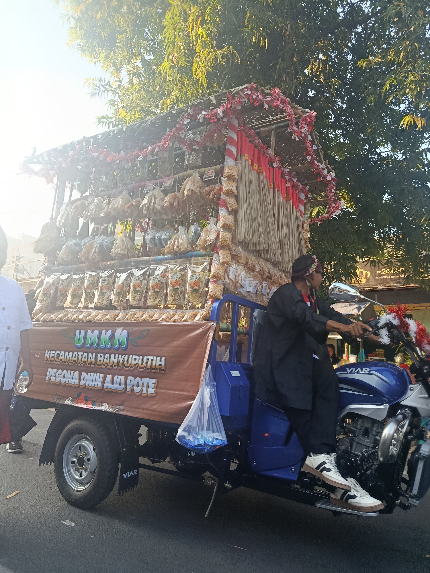 In Ardirejo, Indonesia, a blue and silver Viar three-wheeled motorcycle is being driven by a person wearing black clothing and a head covering. The motorcycle is decorated with red and white garlands. Attached to the rear of the motorcycle is a cart laden with numerous small, packaged food items. A large brown banner on the cart displays the text "UMKM KECAMATAN BANYUPUTIH PESONA DHIN AJU POTE" in yellow lettering. Another person in a white shirt stands to the left of the cart. The scene is illuminated by sunlight filtering through the leaves of trees.