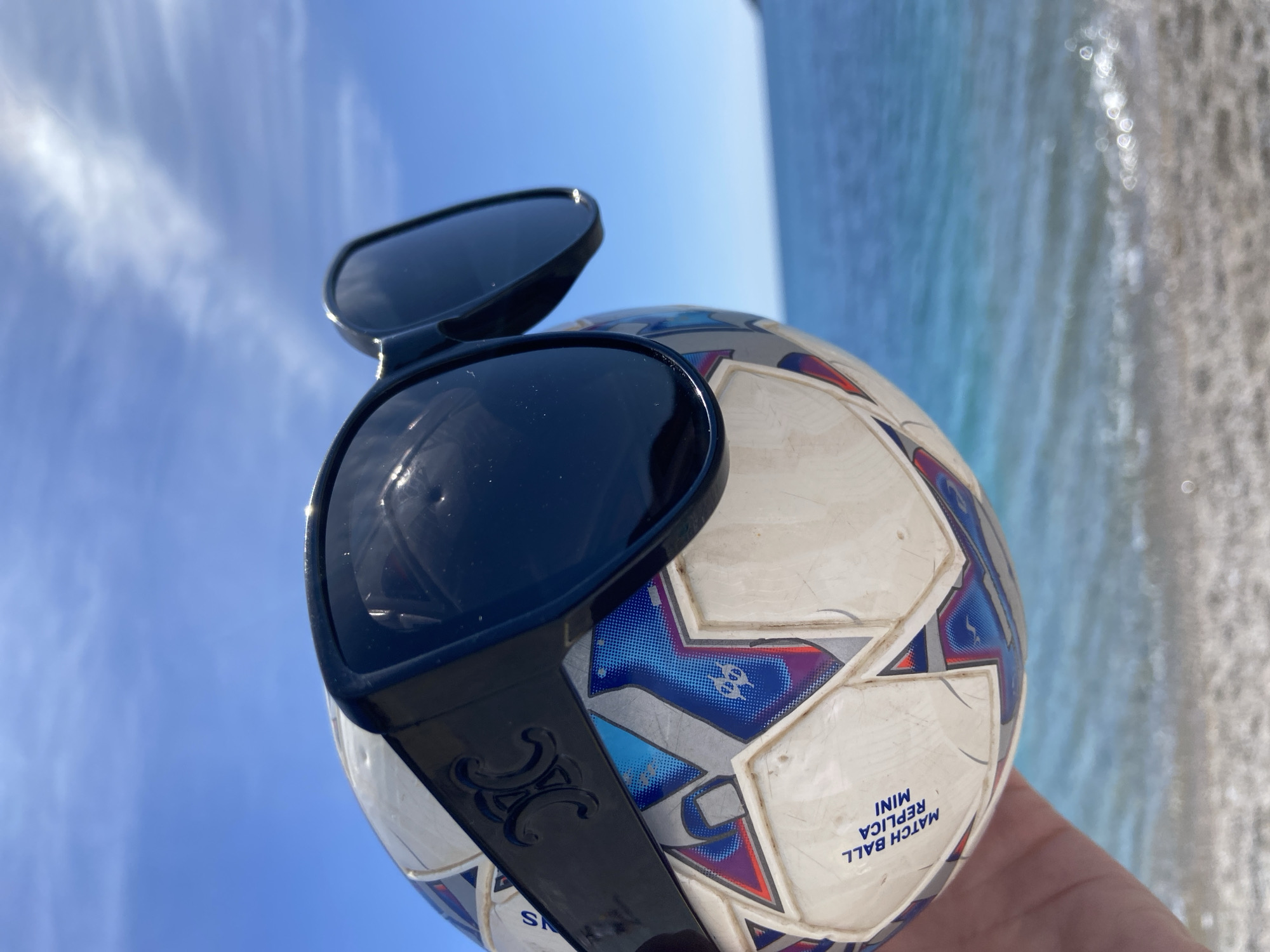A soccer ball with sunglasses is being held up against a backdrop of blue sea and sky. The ball is a "MATCH BALL REPLICA MINI" and has blue and red designs. The sunglasses are black with a decorative design on the arm. The image appears to be taken outdoors, possibly near a beach. The location of Prijevor, Montenegro is not evident in the image.