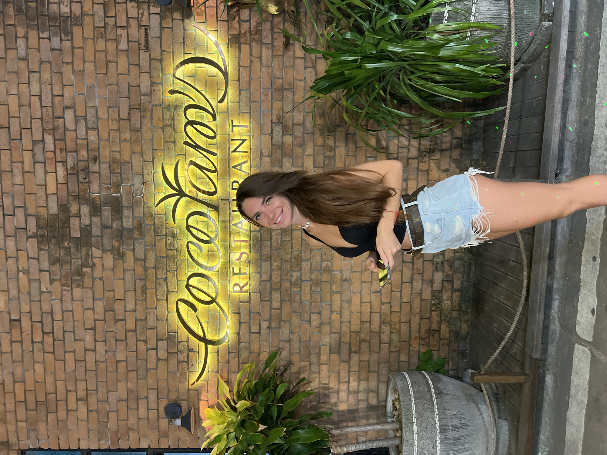 A woman with long, dark brown hair and a wide smile poses in front of a brick wall with a neon sign that reads "Coco Tam's Restaurant" in Baan Bo Phut, Thailand. She is wearing a black top, ripped denim shorts, and a belt with a monogrammed pattern. She is holding a smartphone in her right hand and has a light-colored necklace around her neck. To her left and right are large potted plants, and the ground in front of her is a wooden deck with rope railings. The lighting suggests it is evening.