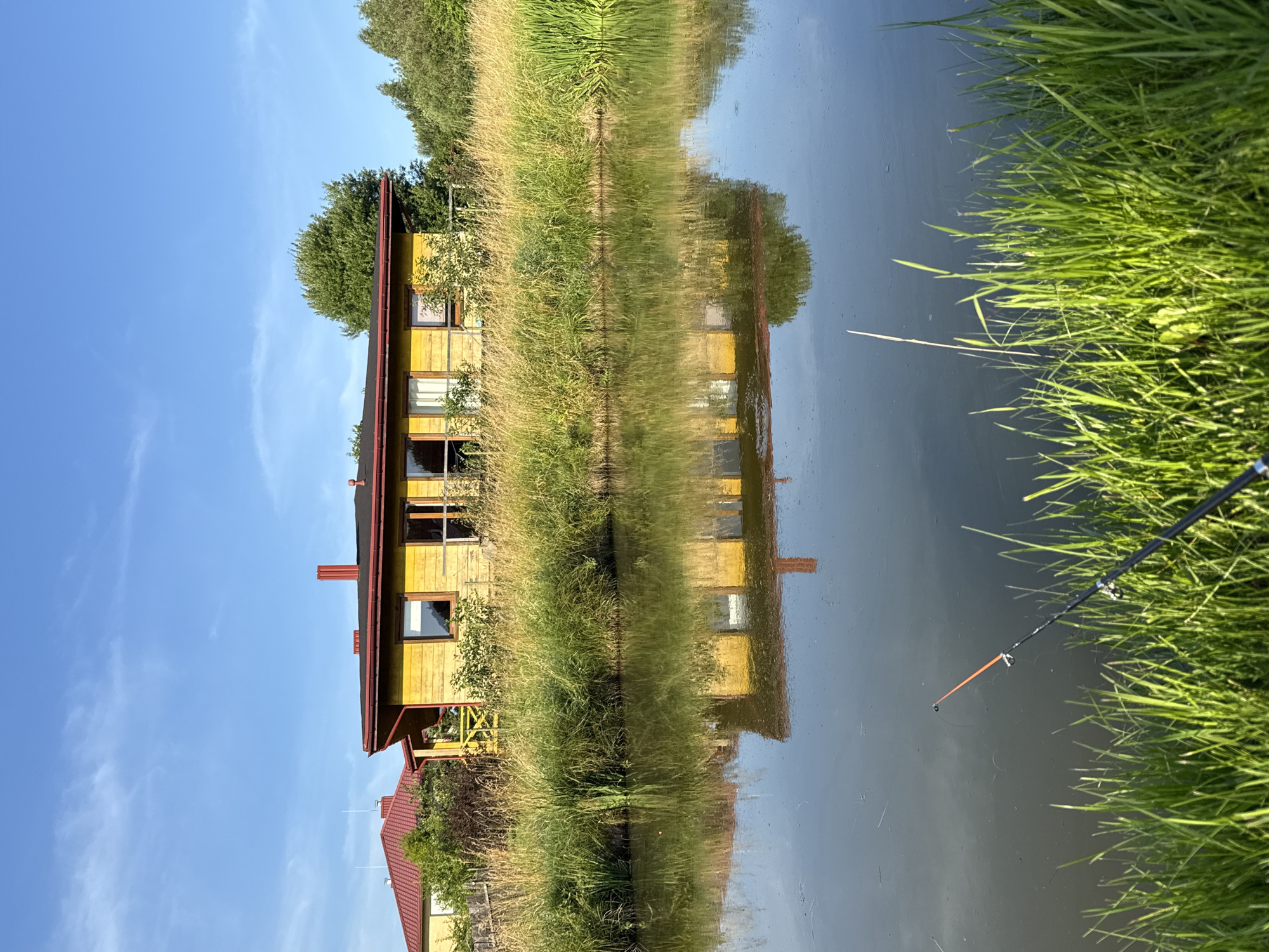 The image depicts a serene waterside scene with a yellow wooden house situated on the bank, surrounded by tall, dry grass. The house features a red roof and a small porch with a yellow railing. The clear blue sky is reflected in the still water, creating a mirror image of the house and surrounding vegetation. In the foreground, a fishing rod rests on the grassy bank, suggesting a peaceful recreational activity. The location is identified as Bendoriai, Lithuania.