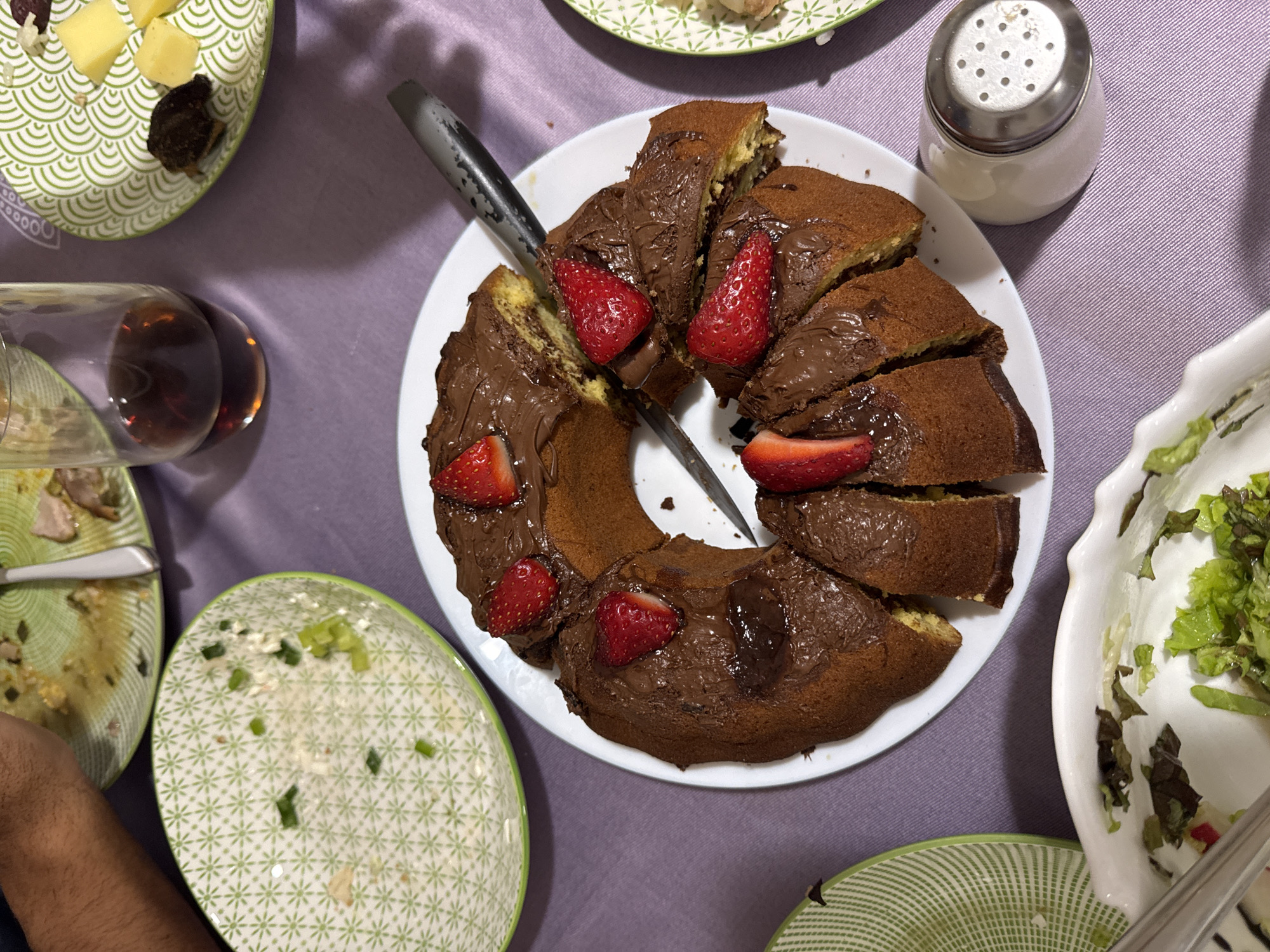 The image shows a table setting with a lavender tablecloth. There's a bundt cake on a white plate, decorated with chocolate spread and sliced strawberries, and a knife placed in the middle. Around the cake are several other plates, some with food remnants, and a bowl of salad. There's also a wine glass with red wine and a salt shaker. The setting appears to be indoors, and the overall arrangement suggests a shared meal or gathering. The location in the city of Burgas, Bulgaria, is not discernible from the image.