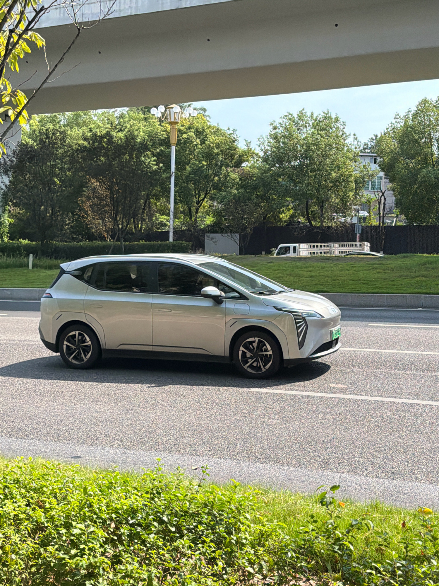 A silver electric vehicle, identified by a green license plate, drives on a paved road under an overpass in Jiangjin, China. The vehicle, a hatchback with a futuristic design, is captured in a profile view, showcasing its aerodynamic lines and distinctive front grille. Sunlight illuminates the scene, casting shadows on the road and the surrounding greenery. The background features lush trees, a grassy embankment, and a black fence. The overpass overhead is a solid concrete structure, with a lamppost visible near its edge. A patch of bright green bushes occupies the foreground, adding a natural element to the urban landscape.