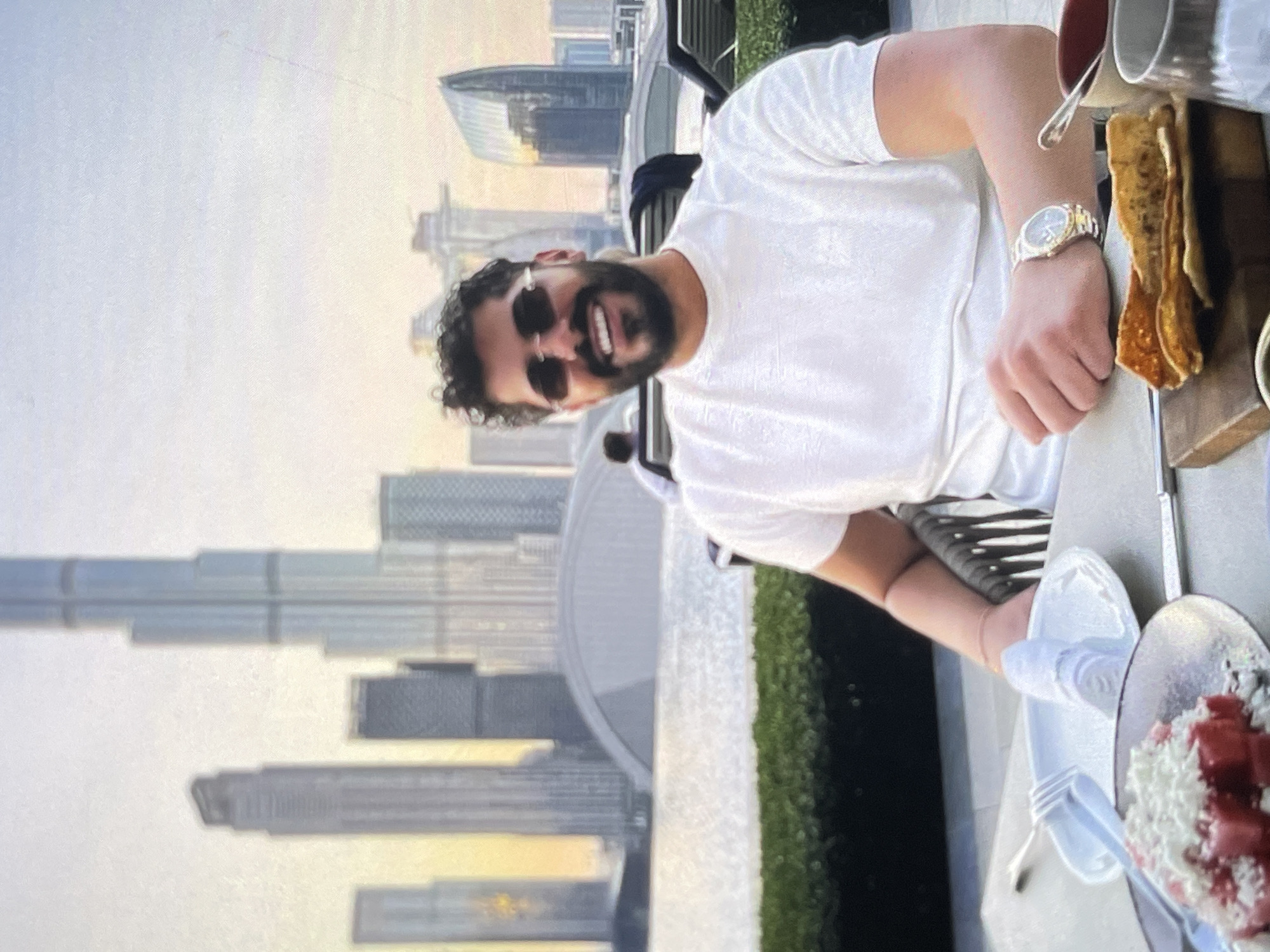 A medium shot captures a man sitting at a table with a city skyline in the background. The man, with dark curly hair and a beard, wears sunglasses, a white t-shirt, and a watch on his left wrist. He is smiling and looking towards the camera. In front of him on the table are plates with food, cutlery, and glasses. The background features several tall skyscrapers, a body of water, and some greenery. The sky is overcast with a hint of sunset colors. The overall scene suggests an outdoor dining experience with a scenic view.