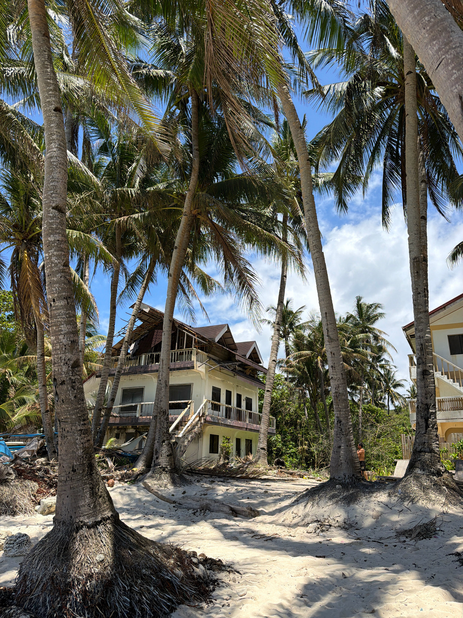 A man stands near palm trees on a sandy beach in Malay, Philippines. A damaged building with its roof caved in is visible behind the trees. The scene appears to be a tropical beach with lush vegetation and blue sky with clouds.