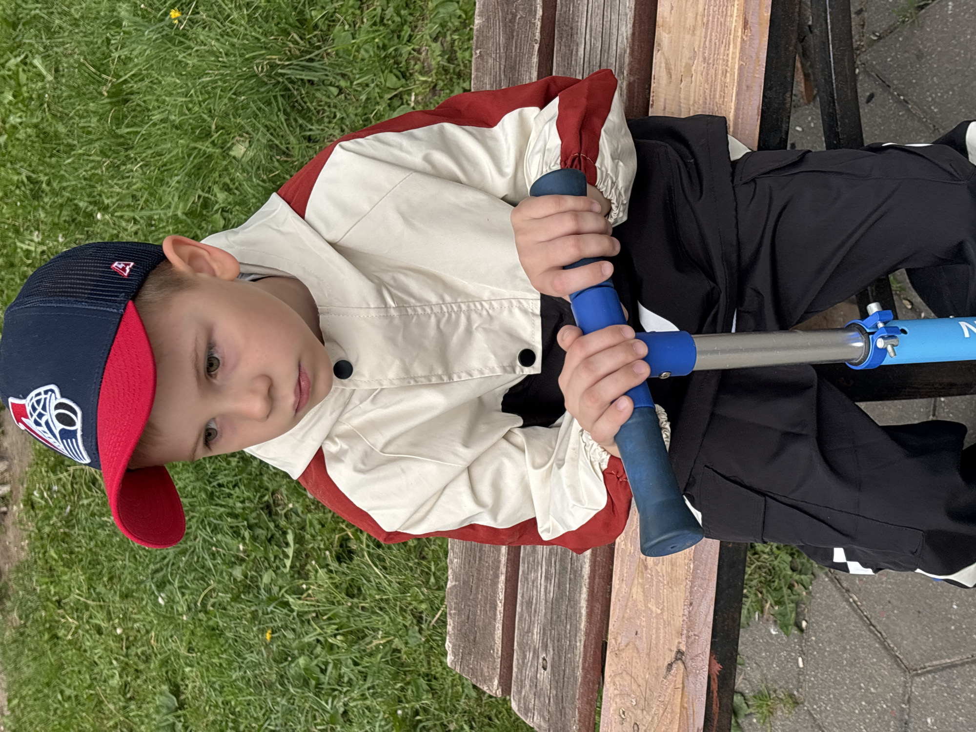 A young boy is seated on a rustic wooden bench outdoors, holding the handlebars of a blue scooter. He is wearing a blue and red baseball cap featuring the "New England Patriots" logo, a cream-colored jacket with red shoulders and black panels, and black pants. His gaze is directed downwards and to his left, giving him a calm and slightly contemplative expression.
The setting appears to be a park or public green space in Rusanovo, Russia. Lush green grass dominates the background to the left, with a small yellow dandelion visible among the blades. The wooden bench shows natural wear and distinct wood grain. To the right of the bench, a section of paved ground, possibly a path or sidewalk, is visible.
The image is captured during the daytime, under soft, natural light, indicating fair weather without harsh shadows. The boy's hands are firmly gripping the blue handles of the scooter, which has a silver metal stem. A partial "N" is visible on the blue stem of the scooter, likely part of a brand name. The scene depicts a quiet moment, perhaps a break from scooter riding, in an outdoor recreational area.