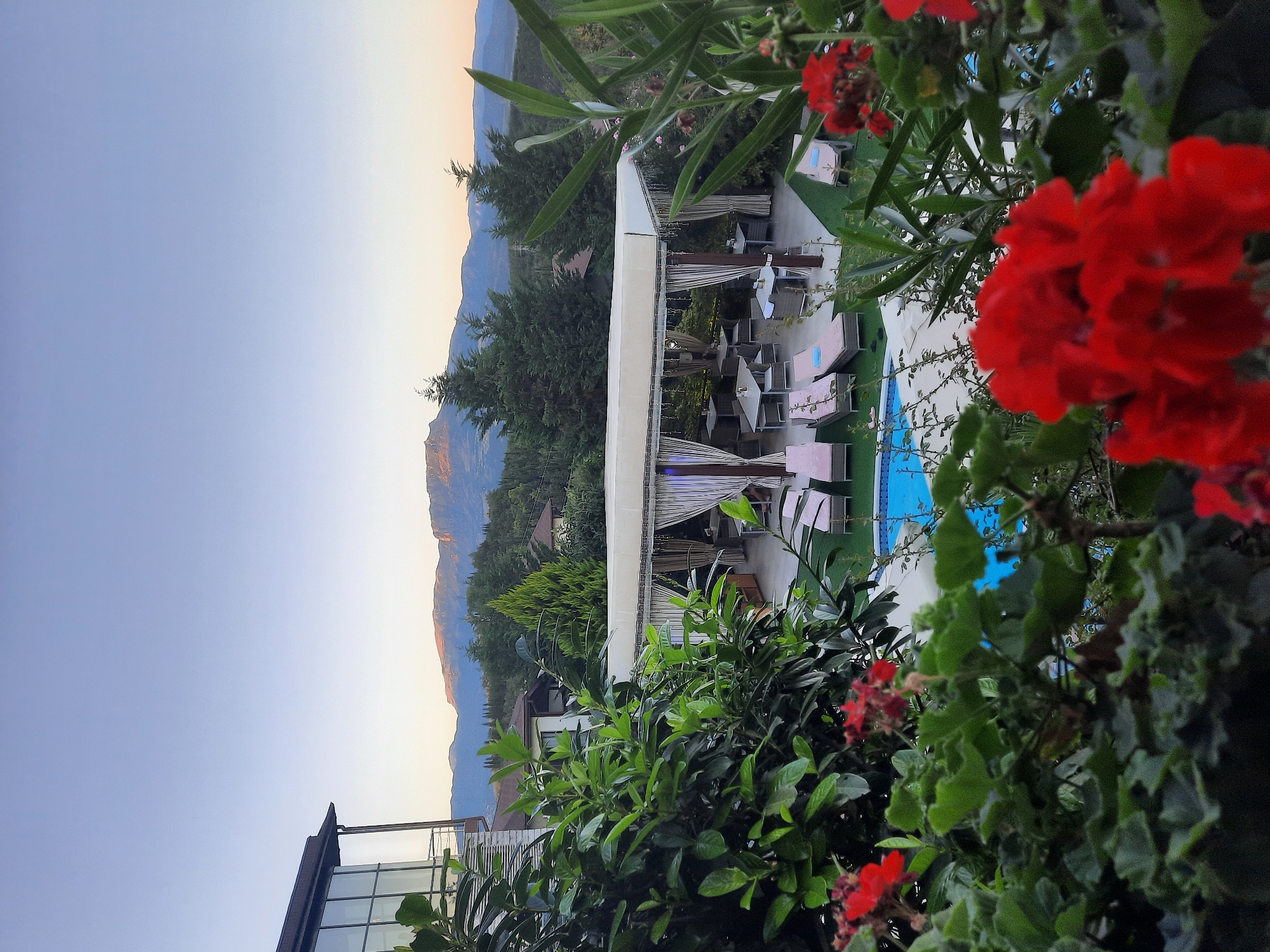 The image depicts an outdoor scene with a swimming pool and lounge chairs, set against a backdrop of mountains. In the foreground, red flowers and green foliage frame the view. A covered patio area with tables and chairs is visible near the pool. A modern building with large windows is situated to the left, and the sky is clear. The scene appears to be a resort or a private residence.