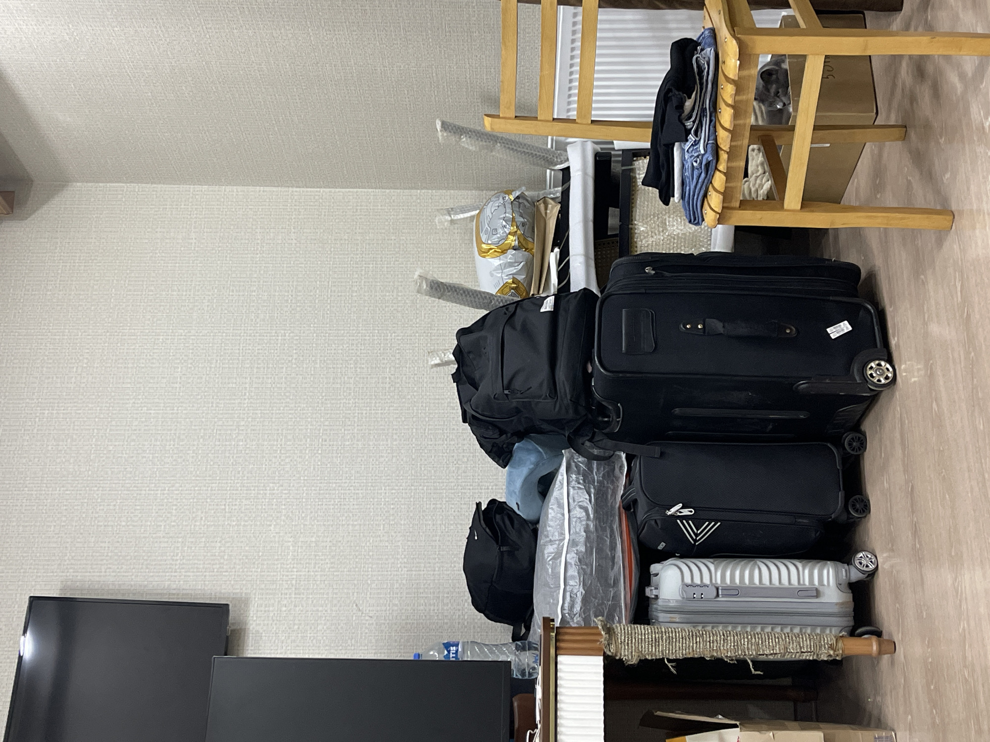 A stack of luggage and bags sits indoors against a light-colored wall. The luggage consists of three suitcases and a backpack, stacked atop a shelf or platform. Additional bags and personal items are packed around the luggage. To the left, part of a television set is visible. A wooden folding chair with clothes draped over it is visible to the right. It's impossible to determine a location within Tbilisi, Georgia based solely on this image.