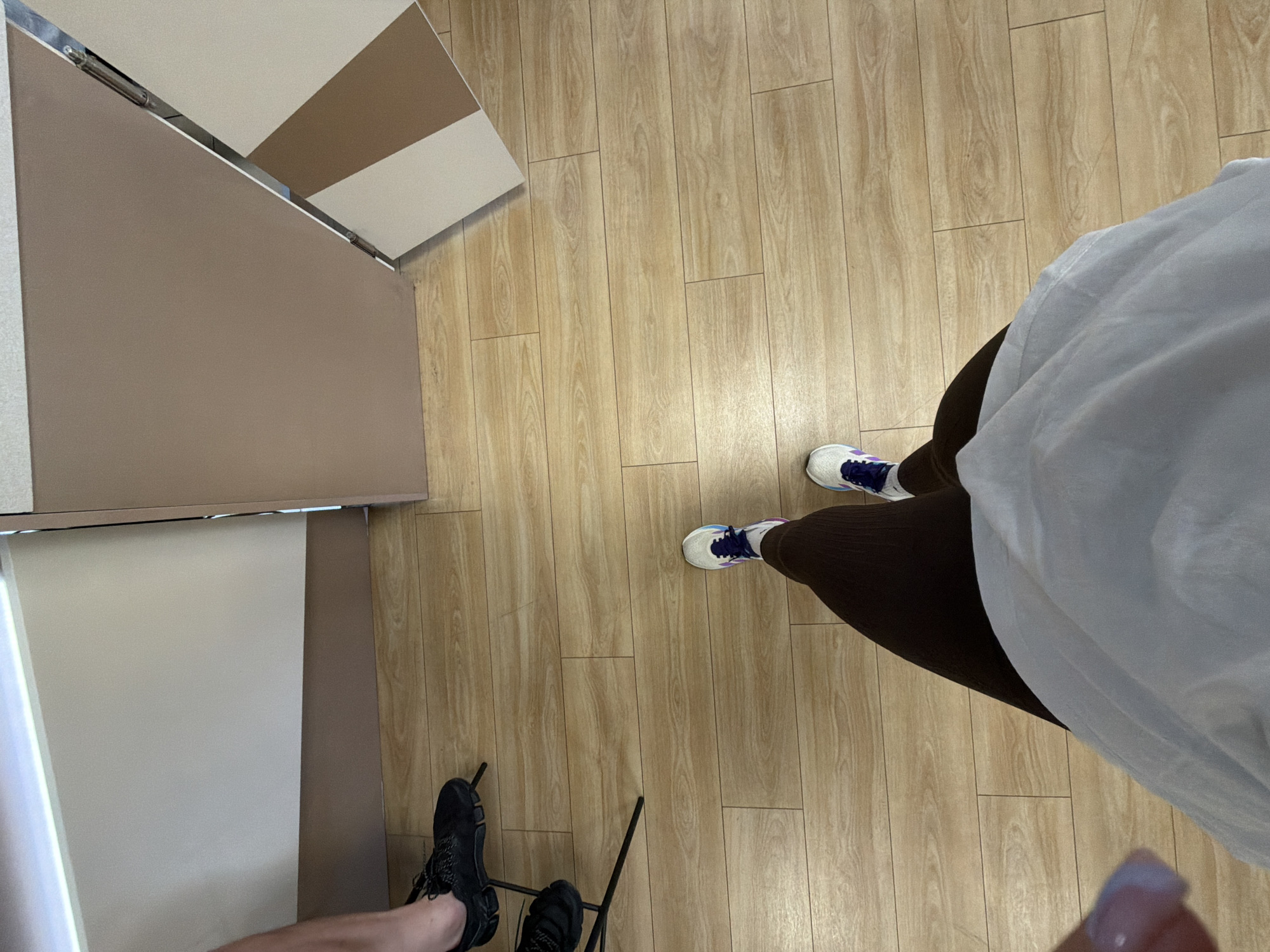 The image shows a first-person perspective looking down onto a light-colored wooden floor. In the upper-left of the frame, part of a wall or furniture is visible, featuring light beige, white, and brown panels. Two pairs of legs and feet are visible. The legs on the right wear dark brown leggings and white sneakers with blue accents. The legs on the left wear black sneakers and are possibly seated on a chair or stool with black metal legs. A white garment partially obscures the person on the right. The image was taken inside an interior space, but there is no evidence to suggest that it was taken in the city of Gabrovo, Bulgaria.