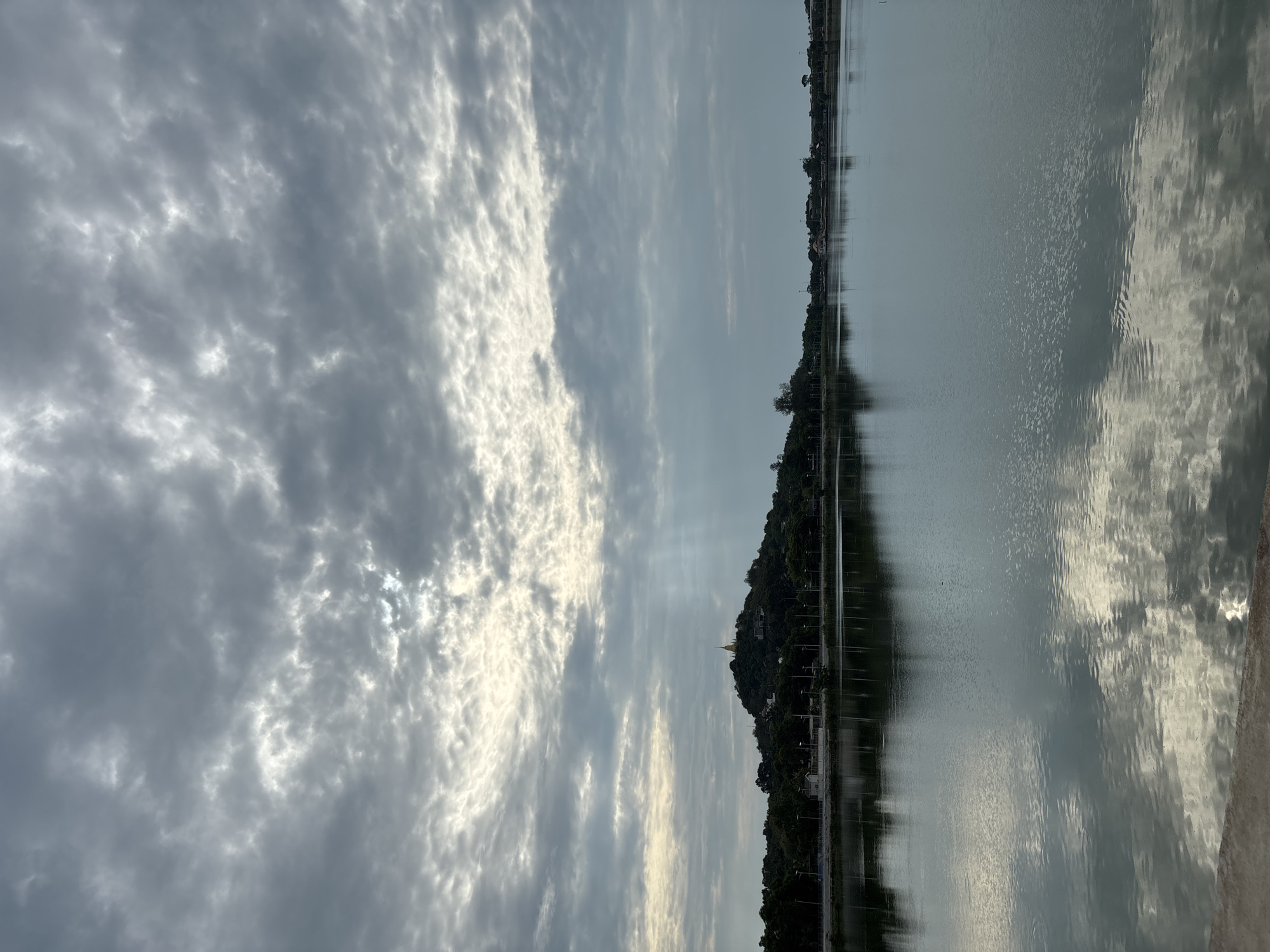 The image depicts a landscape featuring a body of water, potentially a lake or river, reflecting a cloudy sky. The sky is filled with a mix of gray and white clouds, creating a textured and dynamic pattern. In the center of the frame, a small hill or landmass is visible, dotted with trees and what appears to be a golden-colored structure at its peak, likely a temple or pagoda. The water's surface reflects the sky and the landmass, creating a mirrored effect, adding depth to the scene. On the opposite bank of the water, there are faint signs of human presence, such as street lights, houses and a road. Given the reference to บ้านบางรักษ์, Thailand, it is likely that the image captures a serene scene in this region, possibly around a local body of water with a significant religious site. The image suggests a tranquil and reflective atmosphere, with the natural elements dominating the view.