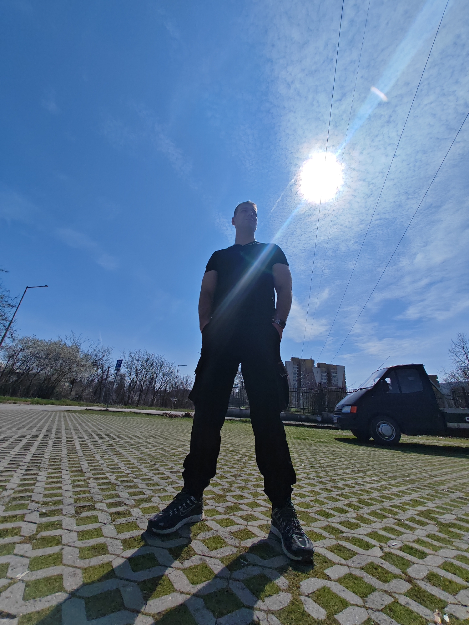 A low-angle, full-body photograph captures a male subject standing on permeable pavers with grass inserts in an outdoor urban environment in Shumen, Bulgaria. The man, positioned centrally, is silhouetted against a bright blue sky with scattered cirrocumulus clouds. He is dressed in a black short-sleeved t-shirt, black cargo pants, and dark athletic shoes (specifically Nike brand on the visible right foot), with a watch on his left wrist. His hands are placed in his pockets, and he gazes upwards and to his left. The sun, situated in the upper right, emits a strong glare and lens flare, illuminating overhead utility lines that traverse the sky. In the mid-ground to the left, a streetlight pole and deciduous trees, some with white blossoms, are visible. To the right of the subject, a dark-colored, compact utility vehicle with a flatbed is parked on the pavers. Behind this vehicle and a metal fence, multi-story residential buildings are present in the background. Distinct shadows are cast on the paver surface from the man and the paver pattern.