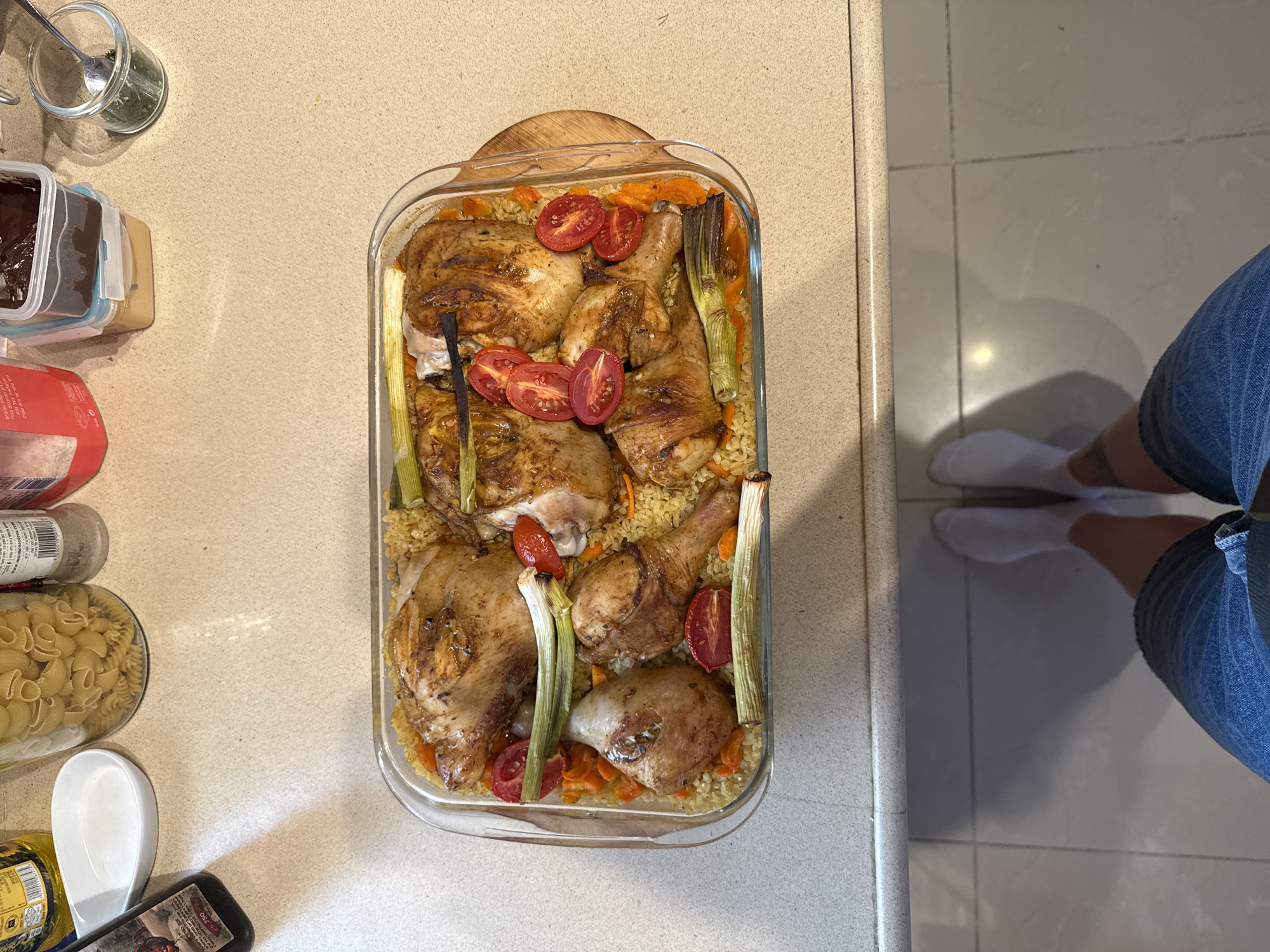 The image depicts a clear glass baking dish filled with cooked chicken pieces, tomatoes, and leeks arranged on a bed of grains, possibly bulgur or couscous, with visible diced carrots. The dish rests on a light-colored countertop. To the left of the dish, there's a collection of kitchen items including jars filled with pasta, herbs, and other food storage containers. A partial view of a person wearing white socks and denim shorts is visible in the lower right corner of the frame. The setting is likely a domestic kitchen. There is no information available that would point to this location being in Rishon LeZion, Israel.