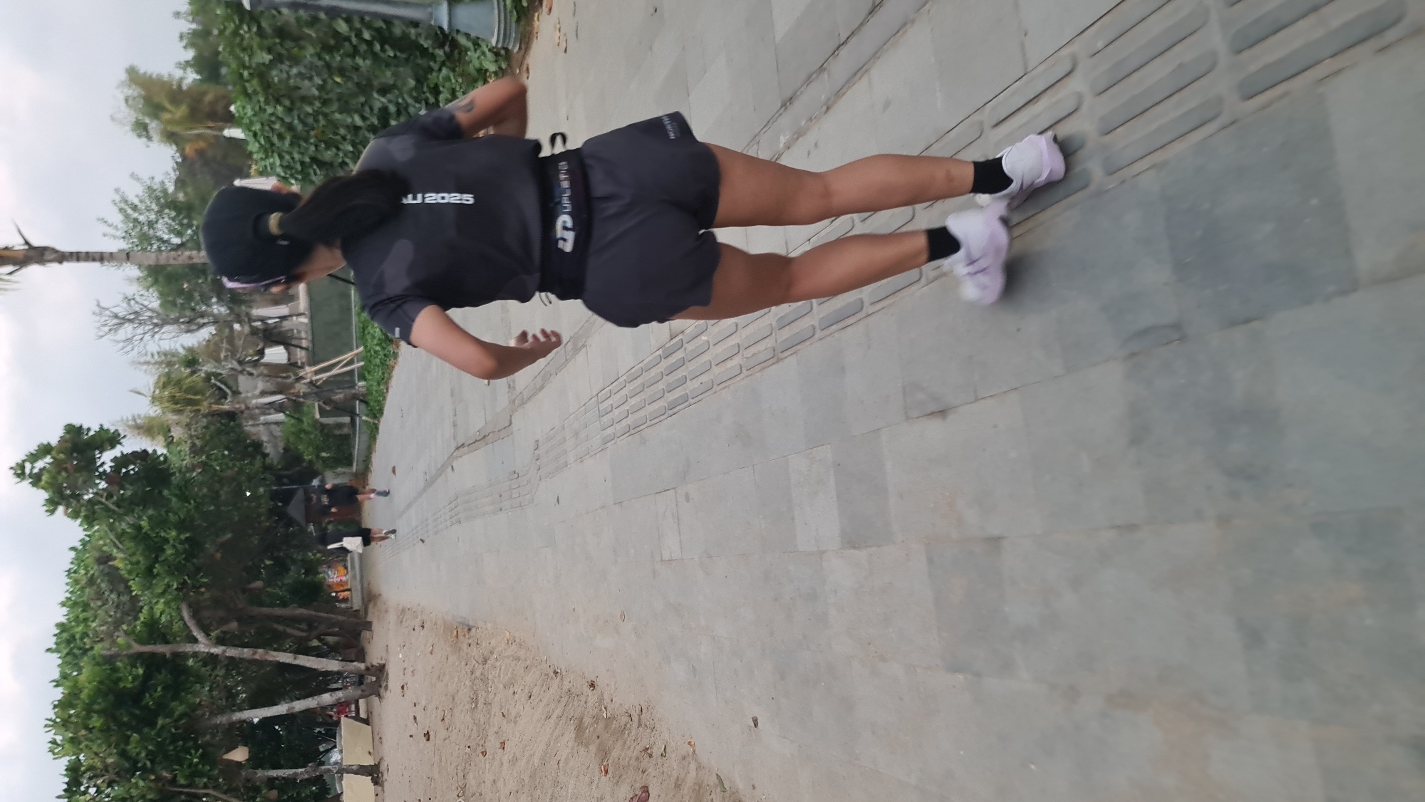 A woman is running on a paved path alongside a sandy beach. The path is lined with textured, raised concrete strips. The woman is wearing a dark t-shirt with white lettering on the back, dark shorts, black socks, and light-colored running shoes. She has a black baseball cap on her head, and her hair is pulled back in a ponytail. She is carrying a black waist pack. In the background, there are several trees with green foliage and a body of water. Two other people are visible in the distance, also running on the path. The scene appears to be in a park or recreational area in Denpasar, Indonesia.