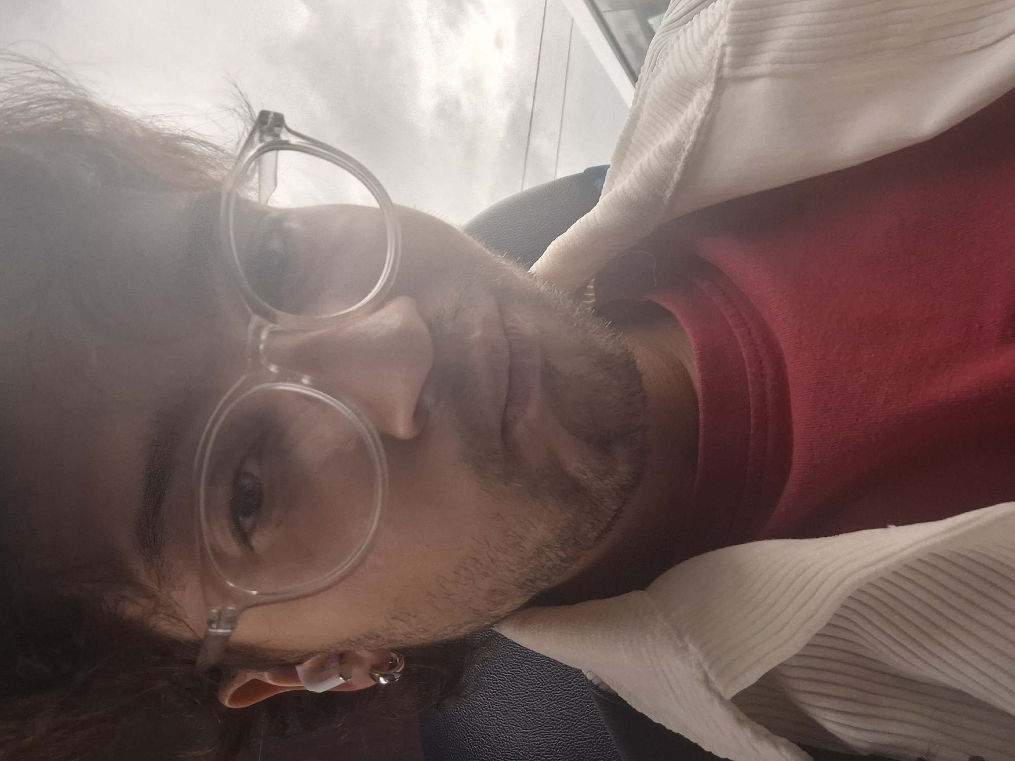 The media file is a close-up, horizontal, selfie-style photo of a person wearing glasses and a red shirt with a white, ribbed cardigan. The person's face is partially obscured by their hair and glasses. They have a light beard and appear to be looking towards the camera. Visible accessories include earbuds in one ear and a ring on the other ear. The background is out of focus, showing a cloudy sky and what might be a window frame. The lighting suggests it could be daytime. The overall impression is casual and personal. There is no visible text. The location context is Natal'ino, Russia.