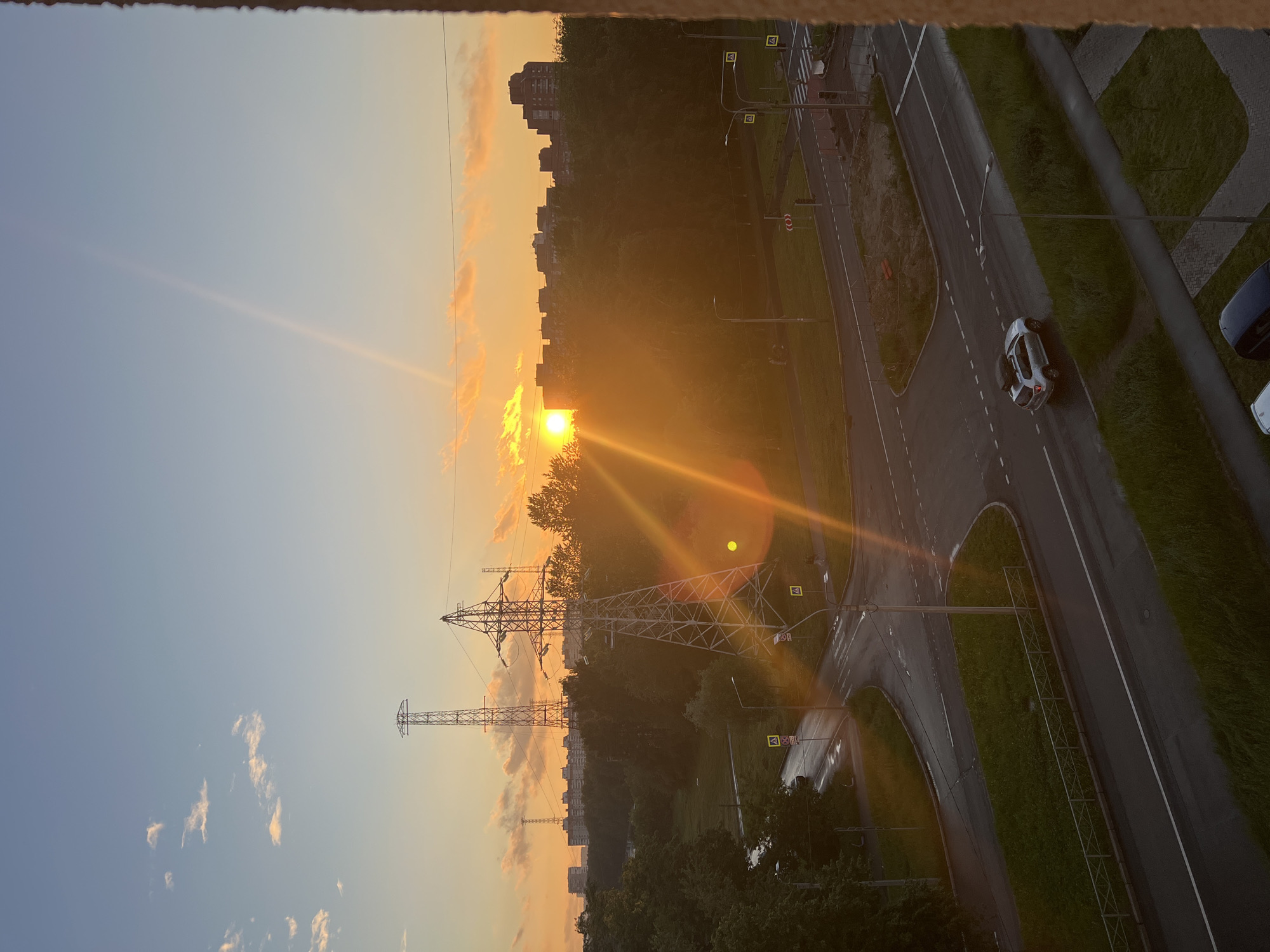 This is a photograph of an urban landscape in Saint Petersburg, Russia, taken during sunset. The sky is a gradient of blue and orange, with the sun setting behind a line of trees and buildings on the horizon. The sun's rays create a strong lens flare effect, casting bright light across the scene.
In the foreground, there is a road intersection with wet pavement, suggesting recent rain. A white SUV is driving on the road. Several tall power line towers are visible in the middle ground, along with a dense treeline. In the background, a cluster of apartment buildings forms the city skyline. The image is taken from an elevated perspective, likely a window of a building.