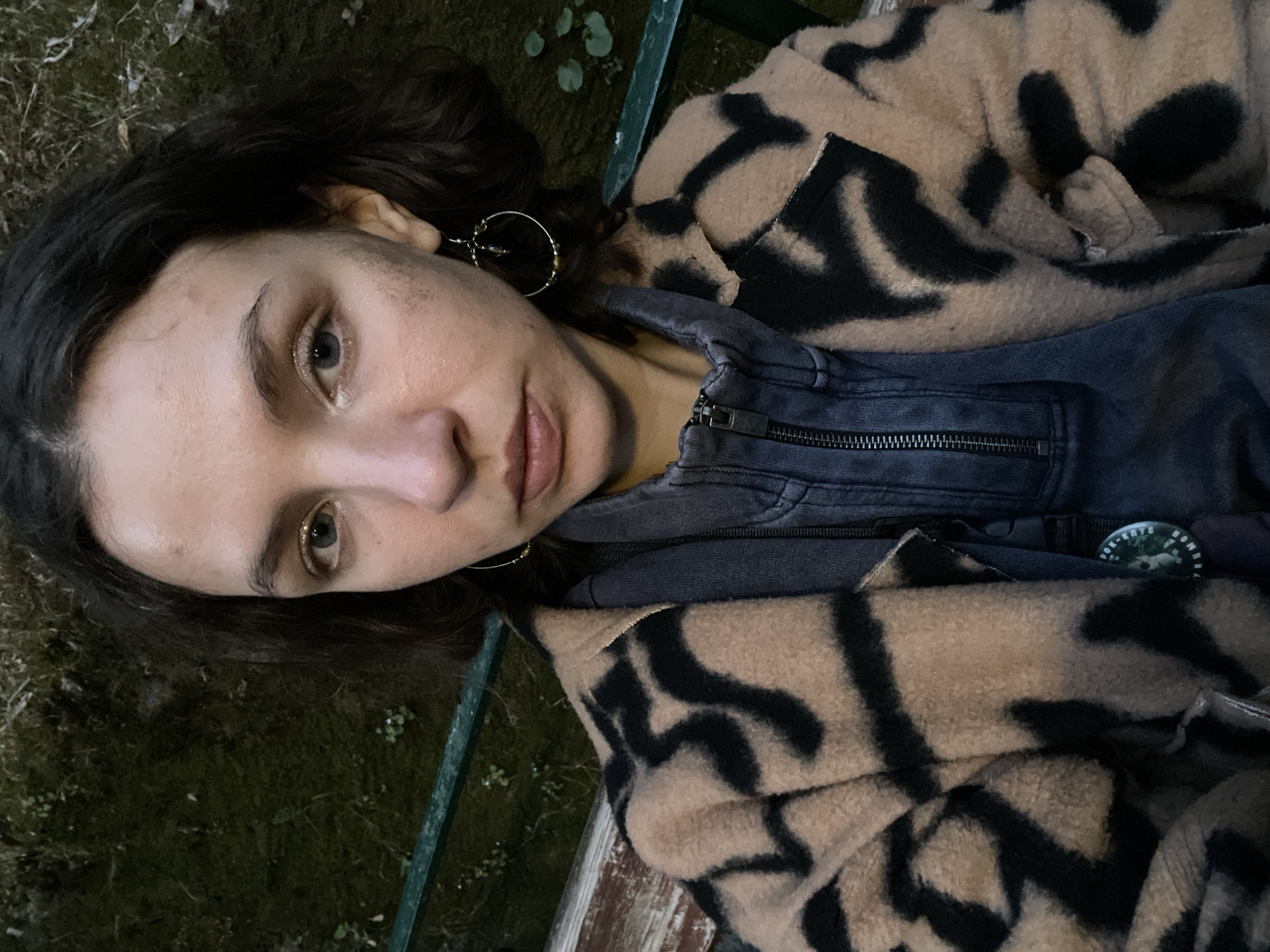 A woman with brown hair and grey eyes is lying on a bench in a park or garden, likely in the Herceg Novi area of Montenegro, given the vegetation. She is wearing a denim jacket under a tan-colored coat with black zebra-like stripes. She has hoop earrings. The area around the bench is dark with some mossy vegetation. The bench has green metal supports.