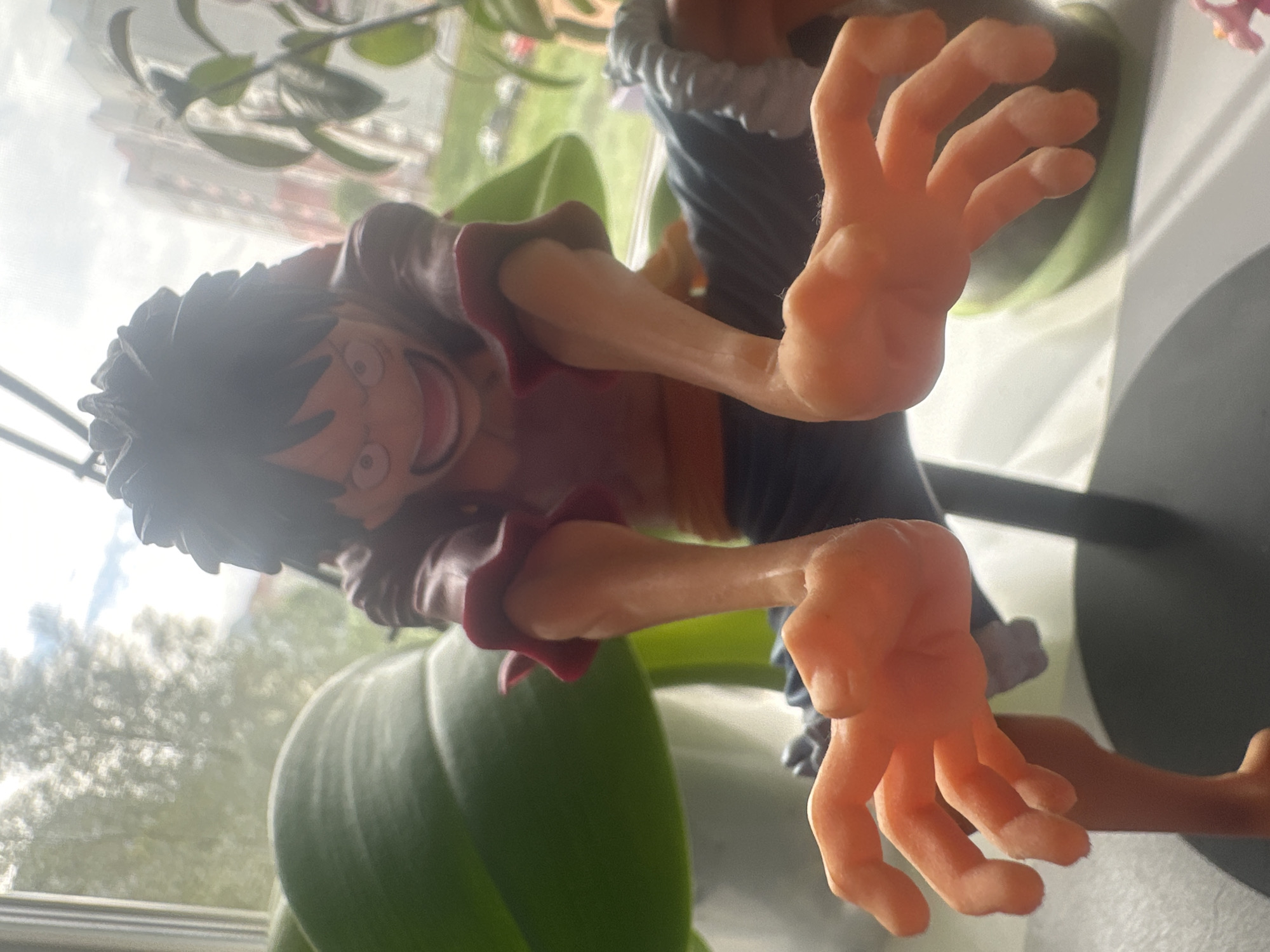 This is a close-up, eye-level shot of a Monkey D. Luffy figurine, positioned in the foreground. The figurine, depicted in a dynamic pose with its arms extended forward and hands open, is the primary subject. Luffy is wearing his signature red shirt and blue shorts. Behind the figurine, a large green leaf of a plant is visible to the left, and a plant with green leaves is to the right. The background shows a blurred view of trees and a residential building, suggesting an outdoor or semi-outdoor setting. The lighting is bright, with a significant amount of light coming from the sky, creating some lens flare. The image appears to be taken indoors, near a window, given the background elements and light source. The exact location in Maladzyechna, Belarus, is not discernible from the image itself.