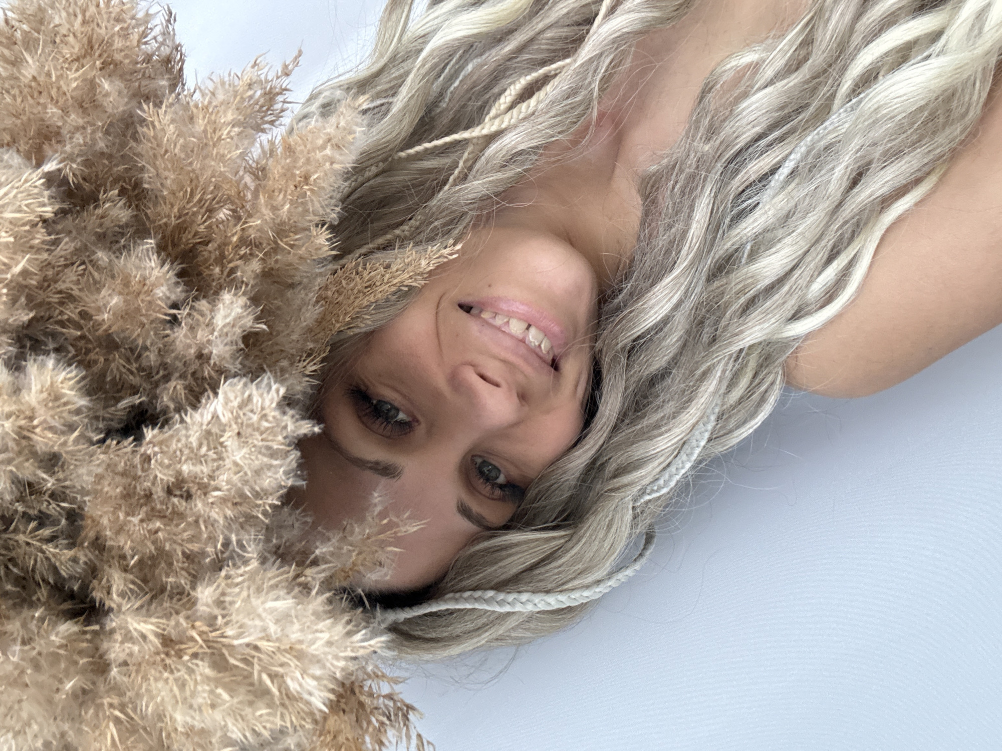 The image depicts a woman with long, wavy blonde hair, some of which is styled into small braids, lying down next to a bundle of dry, light brown reeds or pampas grass. Her face is visible, and she is smiling. The background is a plain, light-colored surface. There is no indication of the location being Rusanovo, Russia, or any specific city.