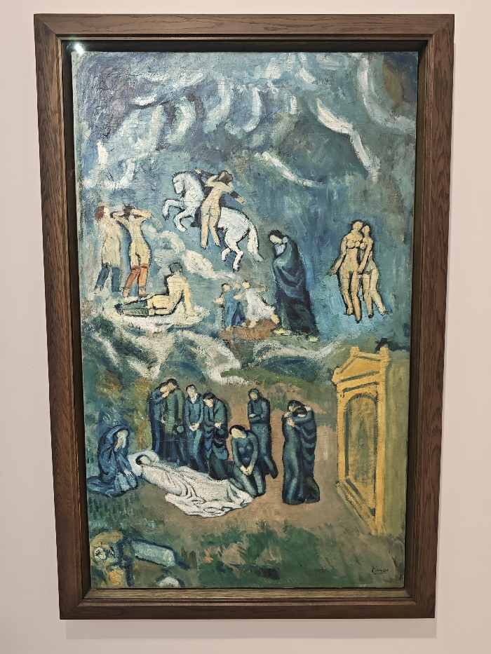 This is a framed painting with somber tones, depicting a funeral scene at the bottom. A body is draped in white, mourned by figures in dark clothing. Above, lighter figures are depicted in a dream-like setting; naked people, couples, and two horsemen. A yellow church or monument stands to the right. The background is a wash of blue and white. The painting is held within a dark brown wooden frame.