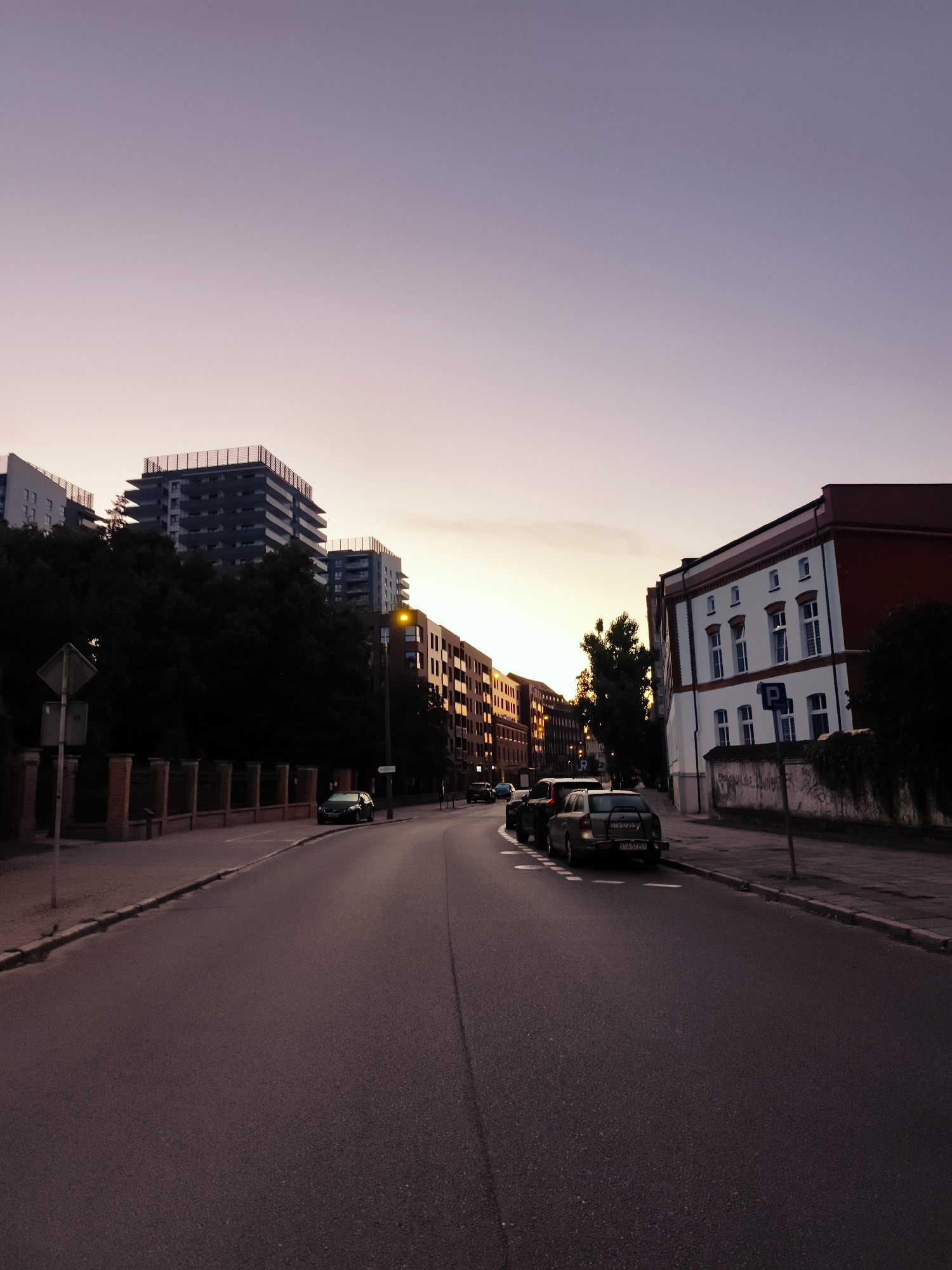 The image depicts a street scene in Gdańsk, Poland, during twilight. The sky is a gradient of soft purple and pink, indicating the sunset or sunrise. On the left side of the street, there is a low brick fence bordering a sidewalk and a line of dark trees. Behind the trees, modern apartment buildings rise, characterized by their flat roofs and balconies. A street lamp with a yellow light is visible further down the road.
On the right side of the street, a building with a red roof and white facade with red trim stands prominently. The building has several windows and appears to be an older structure, possibly with graffiti on the lower wall. Further down the street, more modern buildings with illuminated windows reflect the warm hues of the sky. Several cars are parked along the street or driving. In the foreground, the asphalt road curves gently to the right. A parked silver station wagon with a license plate visible as "STA 97251" is in the middle ground, followed by other vehicles. The overall atmosphere is calm and serene, characteristic of a city at dusk.