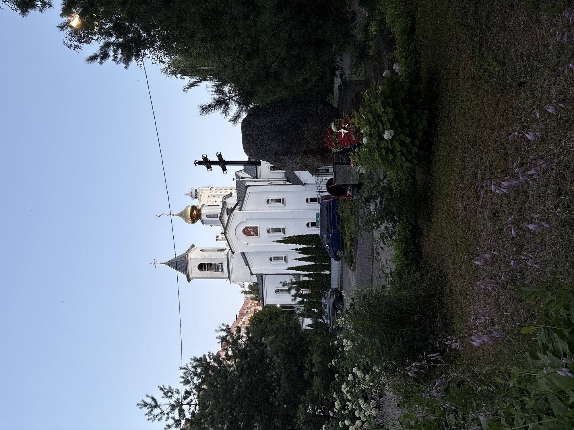 The image depicts the Church of the Holy Assumption of the Blessed Virgin Mary in Vitsebsk, Belarus, located at 5 Pobedy Square. A white church with a golden dome and a bell tower dominates the center of the frame. In front of the church, there is a large dark boulder with a cross mounted on it, appearing to be a memorial or monument. Wreaths of red and white flowers are placed at the base of the boulder. To the left of the church, a tall pine tree stands prominently, and to the right, a dense evergreen tree shades part of the scene. Several other trees and bushes are visible, along with a paved area with two cars parked. The sky above is clear and blue, indicating it is daytime. The overall scene suggests a peaceful and serene atmosphere.